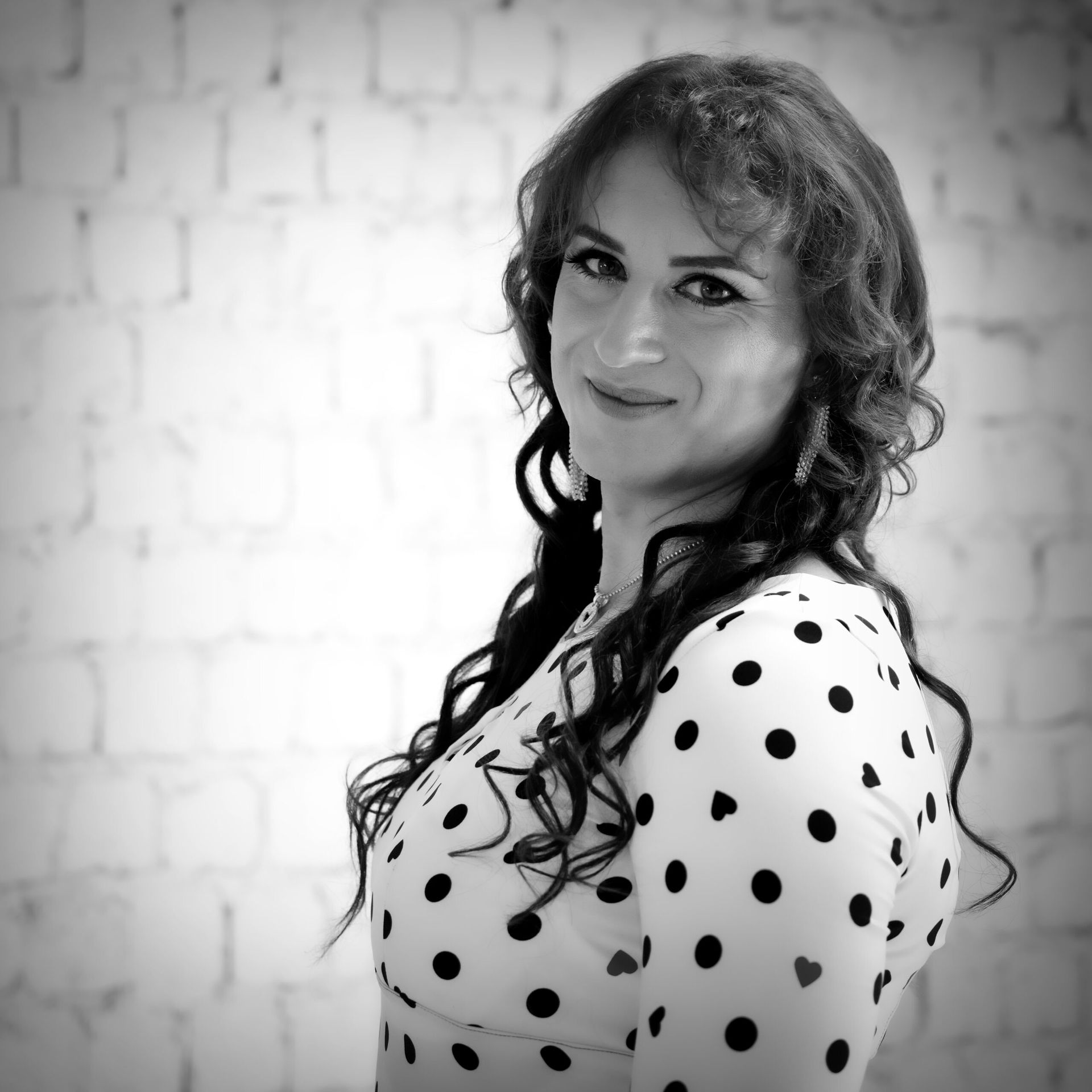 Femme aux longs cheveux bouclés, vêtue d'un haut à pois, souriante, posant devant un mur de briques blanches.