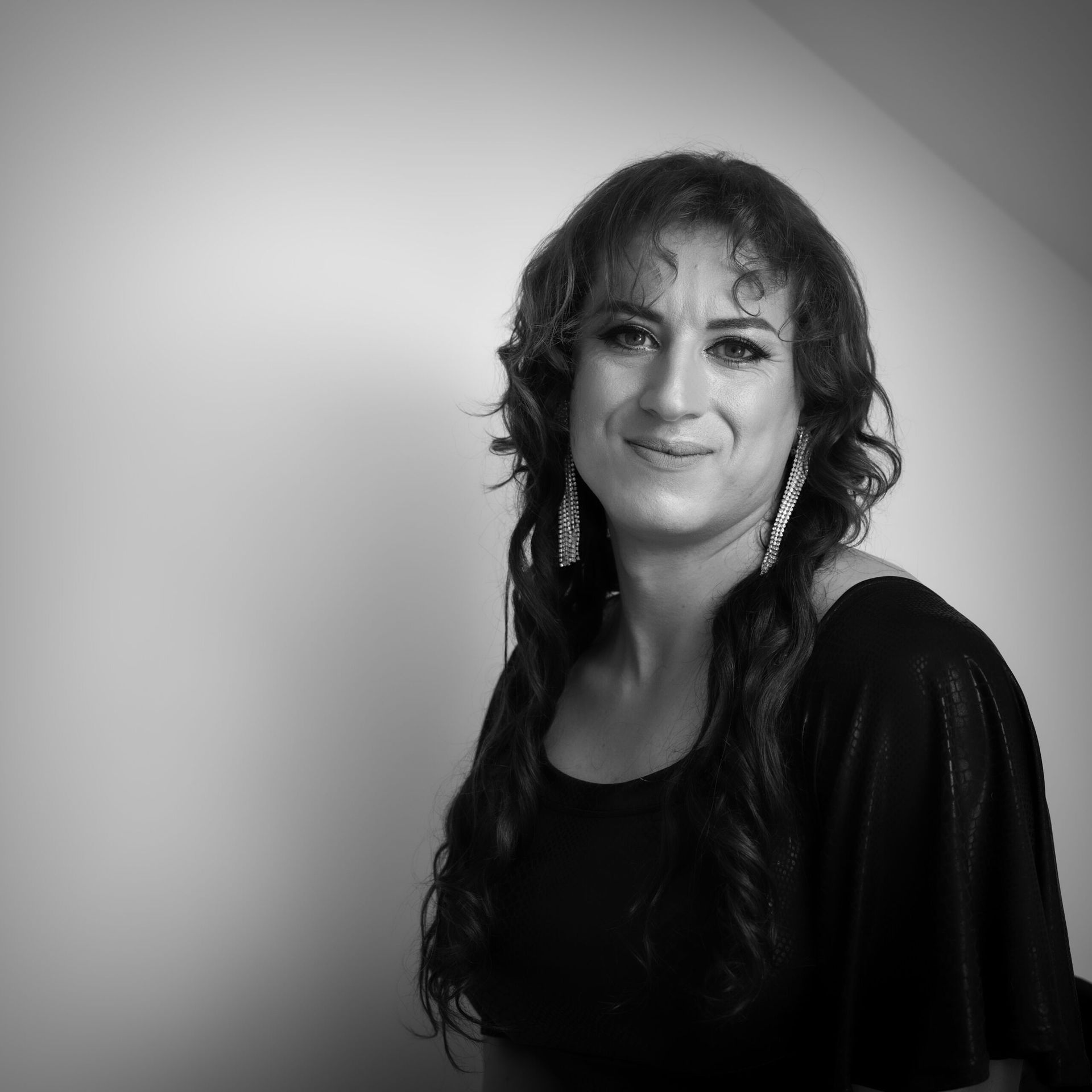 Femme aux longs cheveux ondulés, vêtue d'un haut noir et de boucles d'oreilles pendantes, souriant dans un studio.