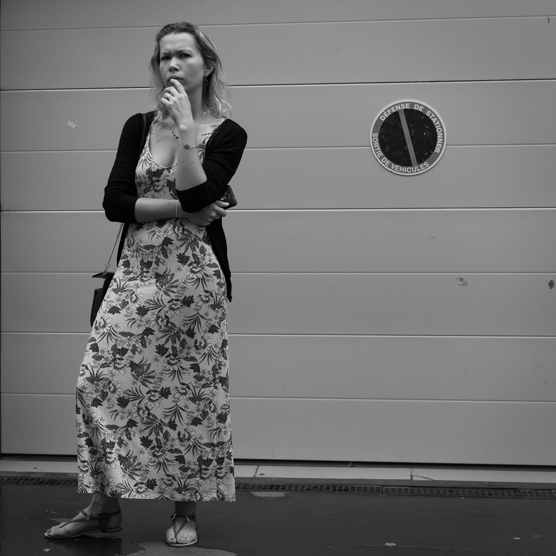 Une femme en robe à fleurs, pensive, se tient debout à côté d'une porte de garage fermée avec un panneau « Stationnement interdit ».