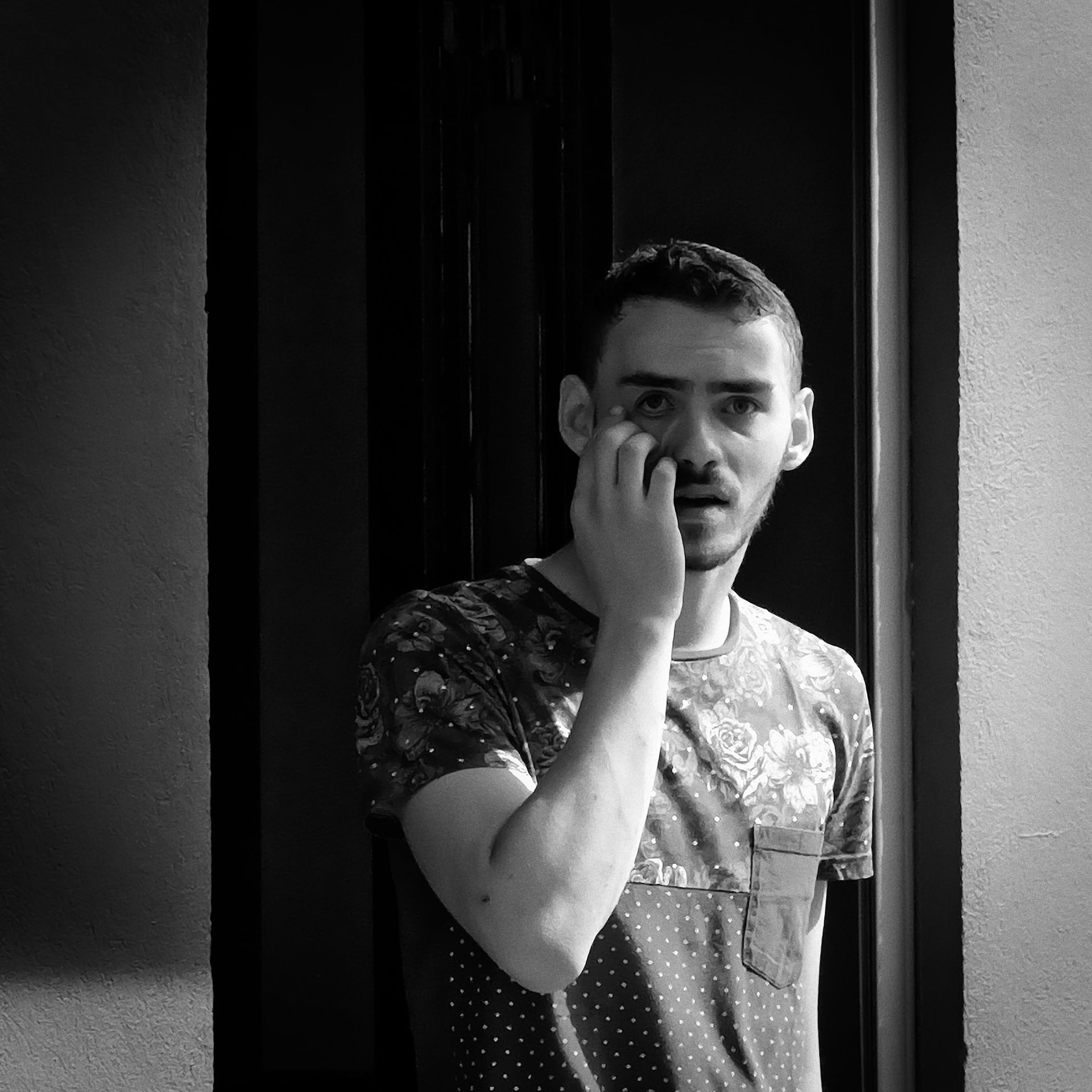 Un homme appuyé contre un chambranle sombre, se touchant le visage, l'air surpris.