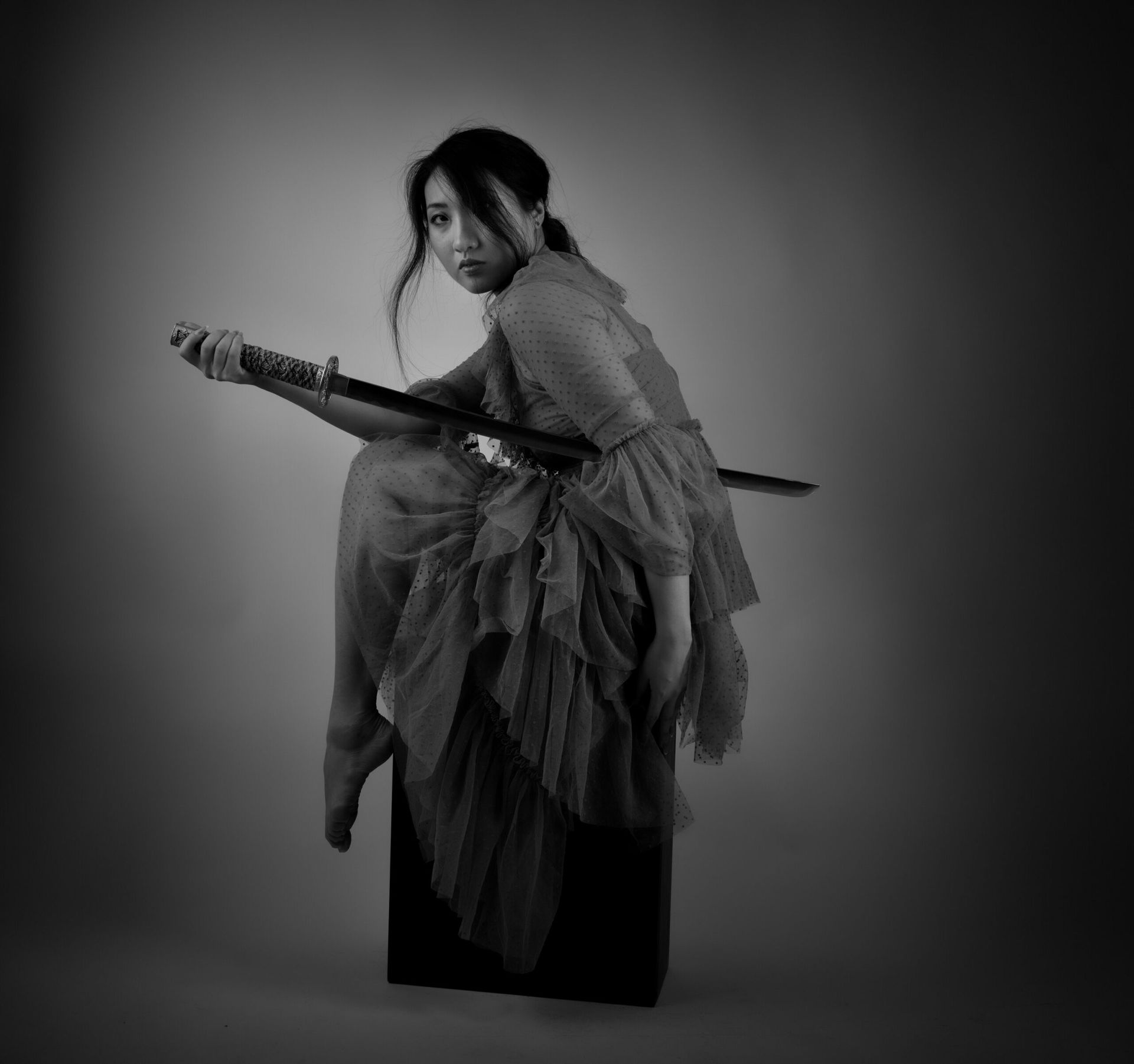 Une femme en robe à volants, tenant une épée, est assise sur une boîte noire.