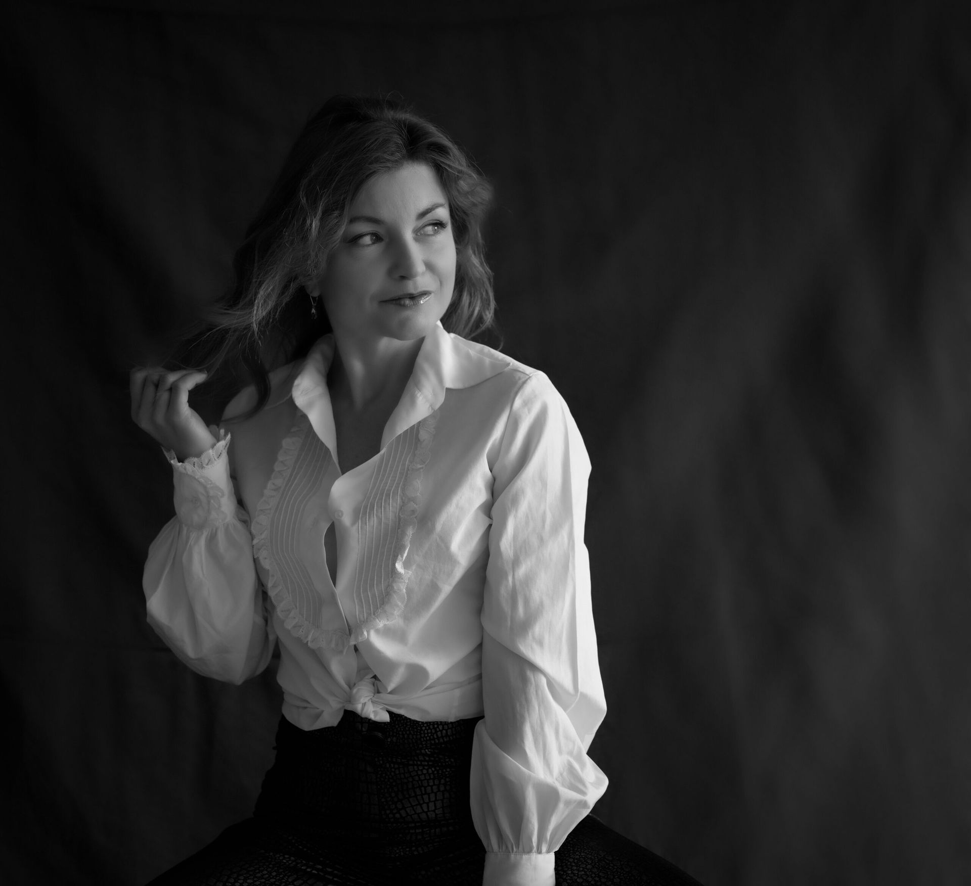 Femme en chemise blanche regardant hors champ, main dans les cheveux, fond noir.