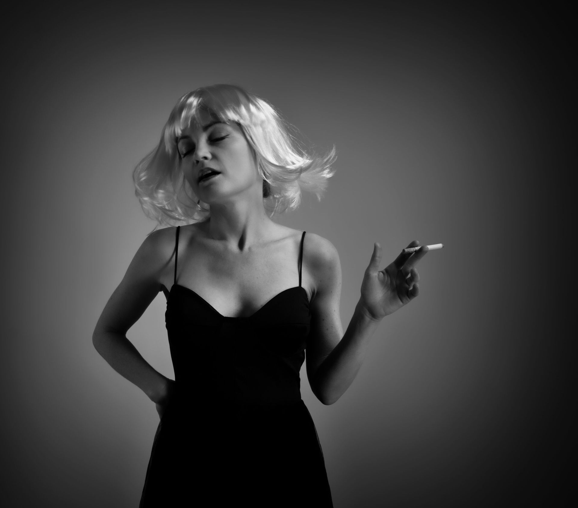 Femme en robe noire et perruque, fumant une cigarette, la main sur la hanche.