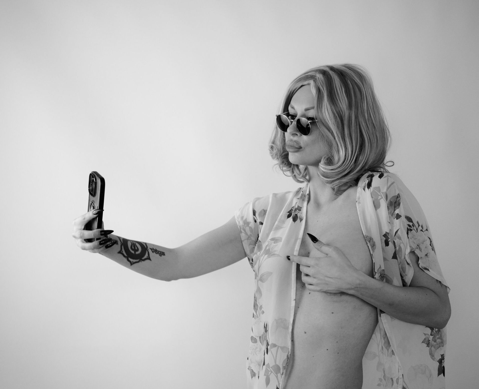 Une personne portant des lunettes de soleil et une perruque prend un selfie avec son téléphone, vêtue d'une robe à fleurs et a un tatouage.
