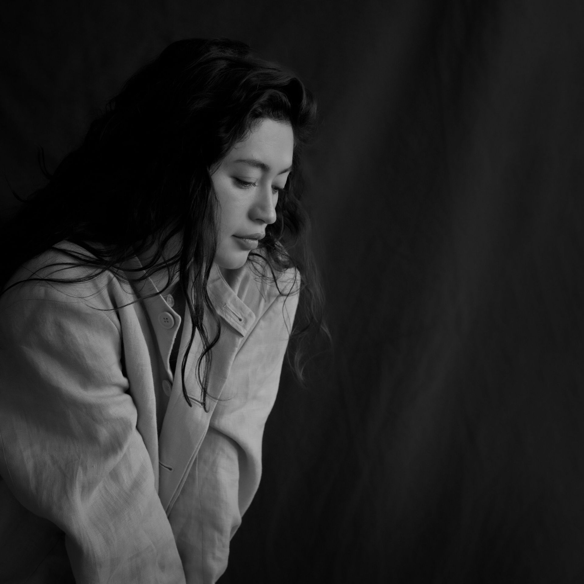 Femme aux longs cheveux noirs regardant vers le bas, portant une veste claire, sur fond noir.