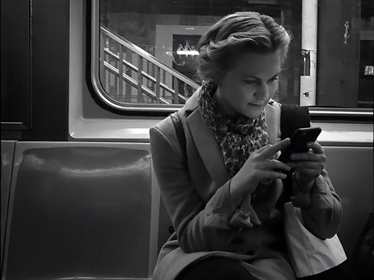 Femme dans le métro utilisant un smartphone, assise près d'une fenêtre, portant une écharpe, en noir et blanc.