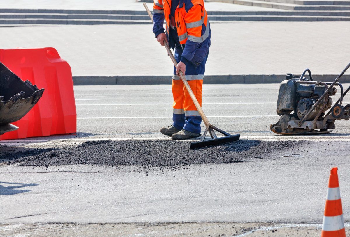 A Man In Orange Uniform Is Repairing A Pothole In The Road — Elite Bitumen & Asphalt In Mackay, QLD