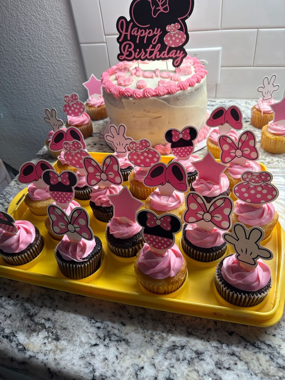 Pink birthday cake and cupcakes topped with butterfly decorations on a yellow tray in a kitchen