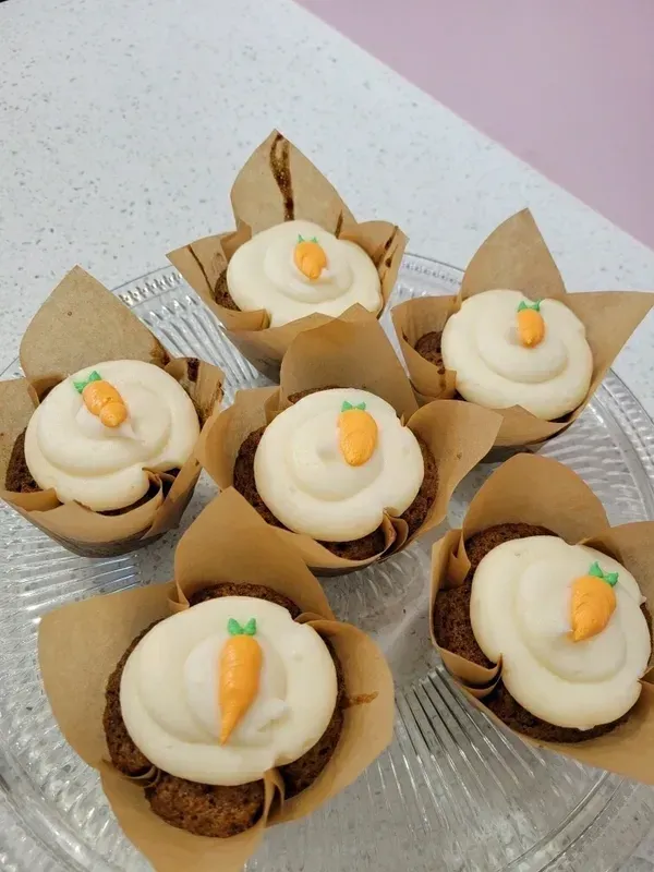Six carrot-topped cupcakes with white frosting in brown liners on a glass plate