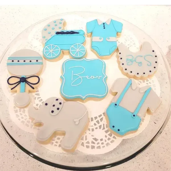 Blue baby-themed decorated cookies on a glass plate, including a onesie, stroller, bib, rattle, and bear.