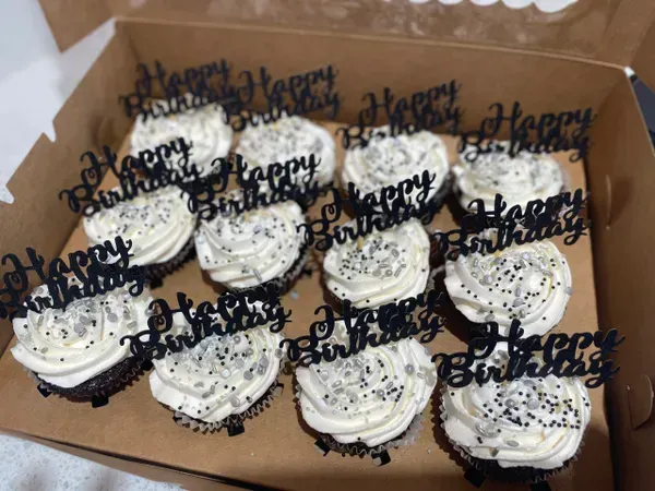 Box of white-frosted cupcakes with black “Happy” toppers and sprinkles