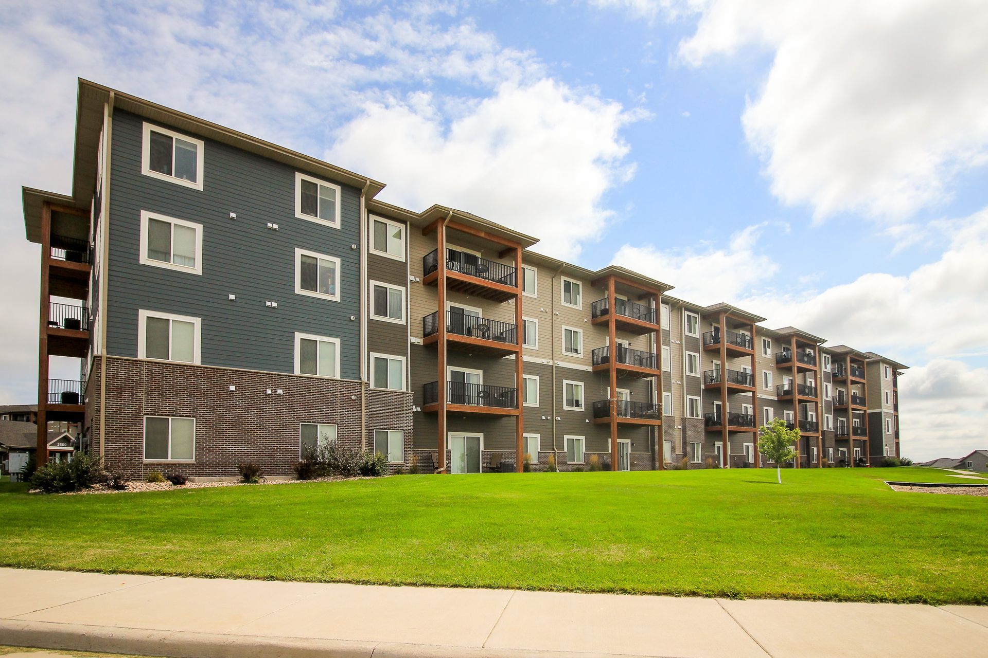 Apartment building with blue and tan siding, multiple floors, balconies, and a green lawn under a cloudy sky.