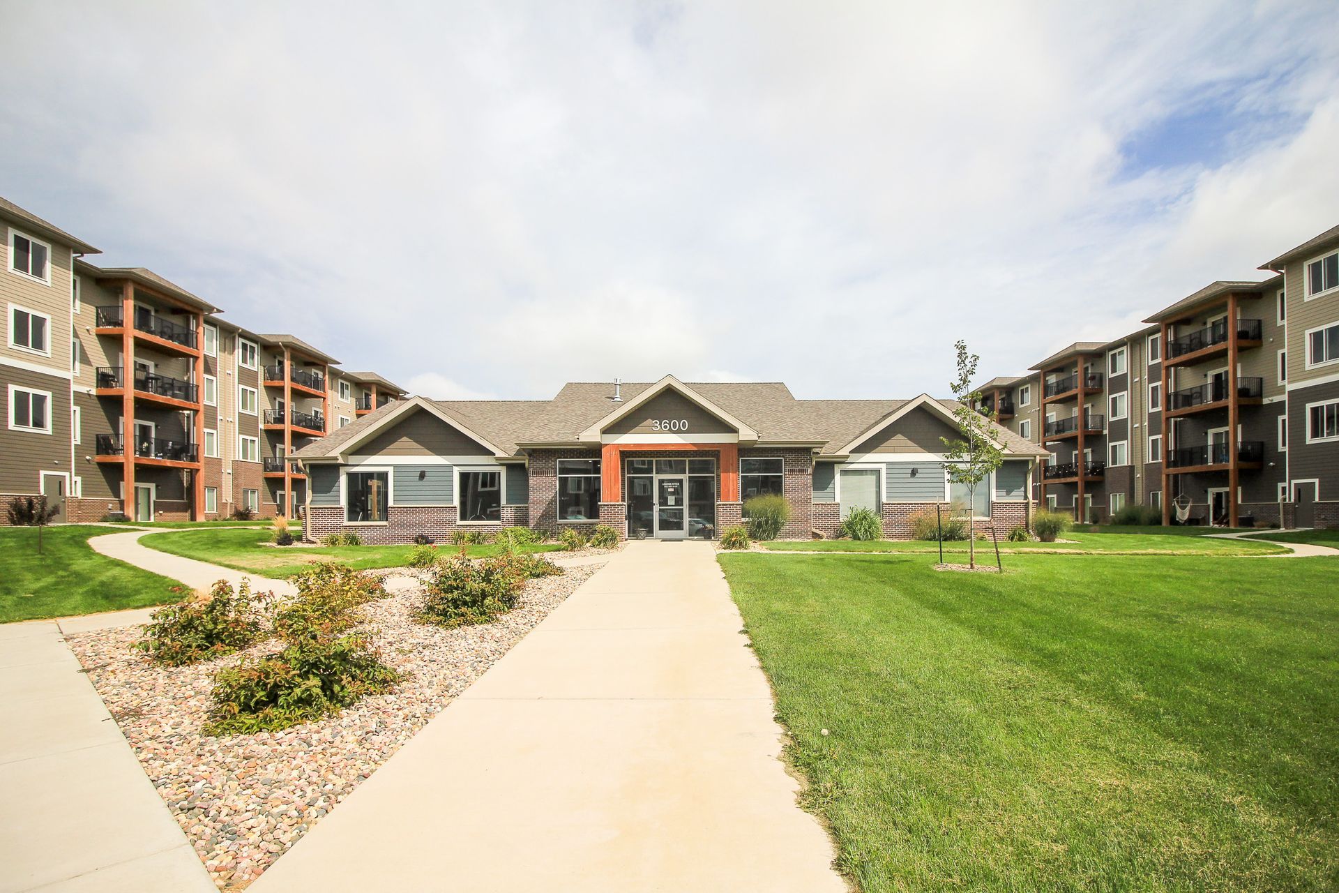 A long, paved path leads to a small building with an open entryway, flanked by multi-story apartment buildings and green lawns under a cloudy sky.