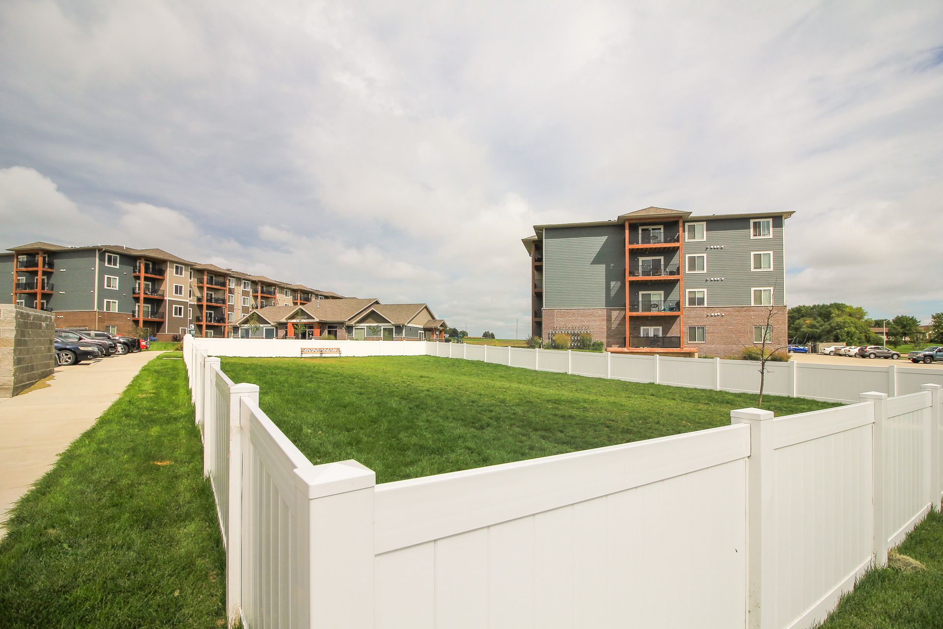 Green fenced-in lawn in front of multi-story apartment buildings under cloudy sky.
