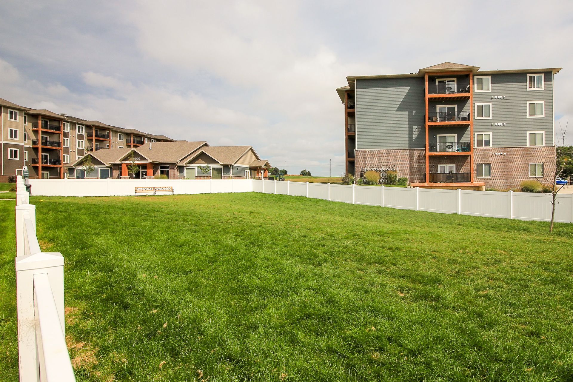 A grassy lot surrounded by a white fence, with multi-story apartment buildings in the background under a cloudy sky.