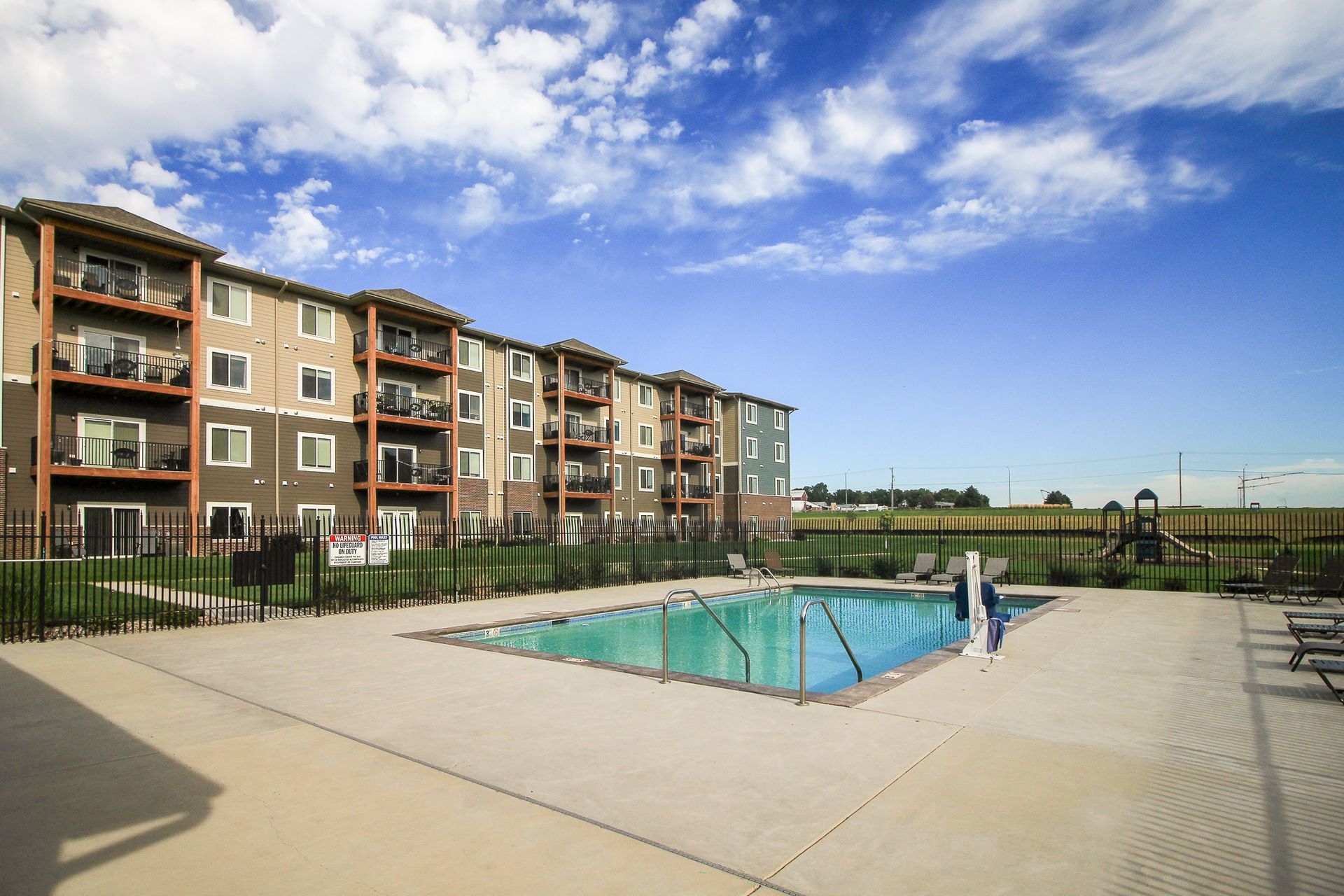 Apartment complex with a pool on a sunny day. Buildings are tan and brown with balconies. The pool is surrounded by concrete.