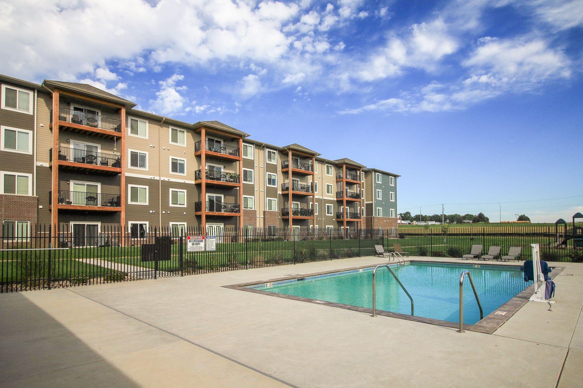Apartment building with a pool on a sunny day. Brown, beige, and blue building, with a pool, lawn, and fence.