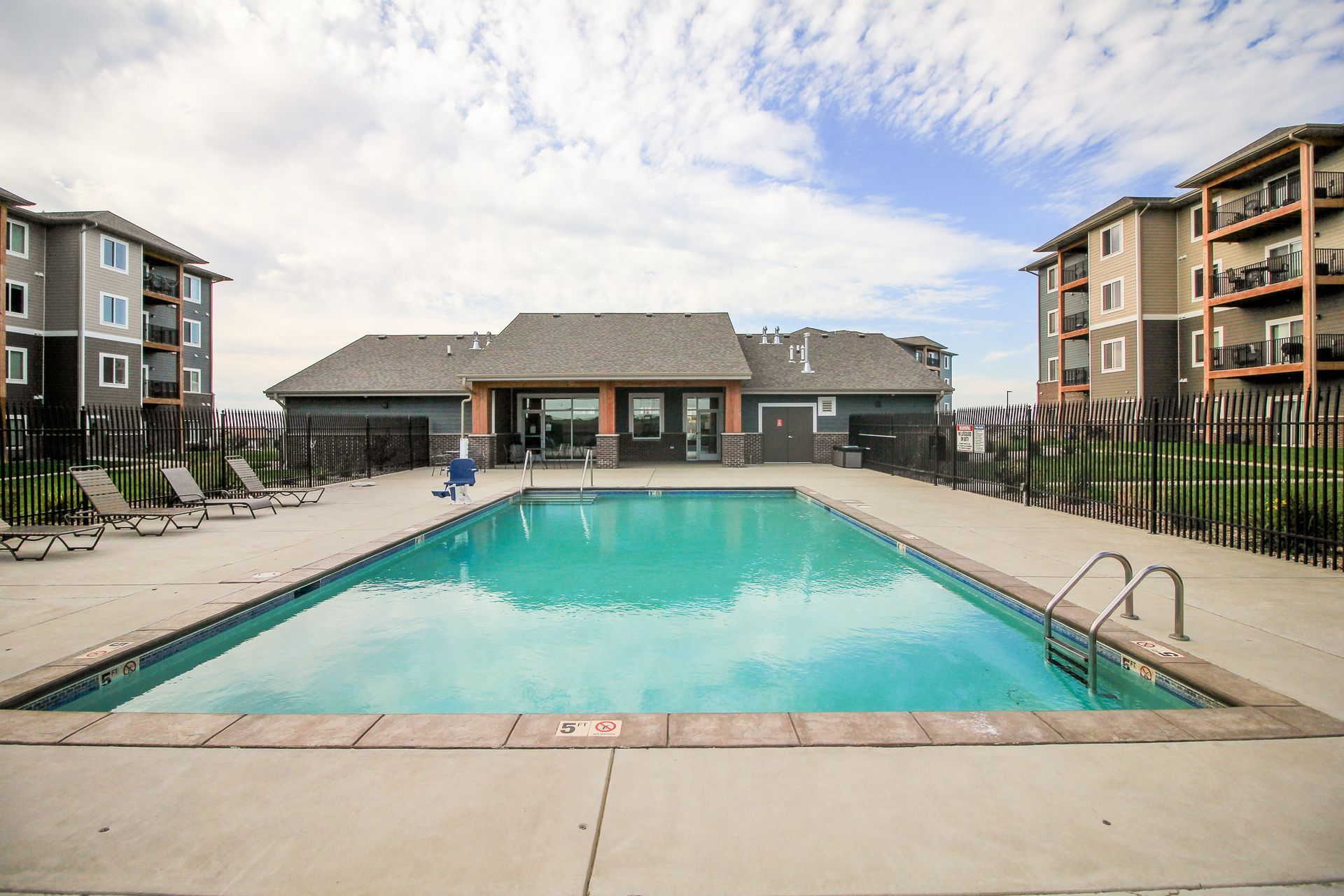 Swimming pool between two apartment buildings on a sunny day. Lounging chairs are visible on the pool deck.