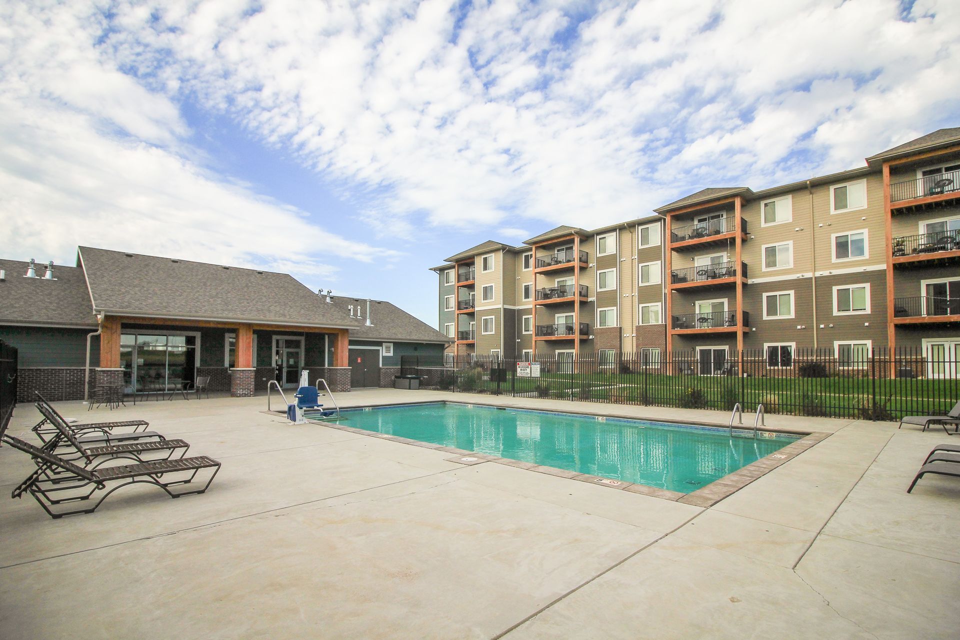 Outdoor pool and patio area at an apartment complex on a sunny day. There are lounge chairs and a building with balconies.