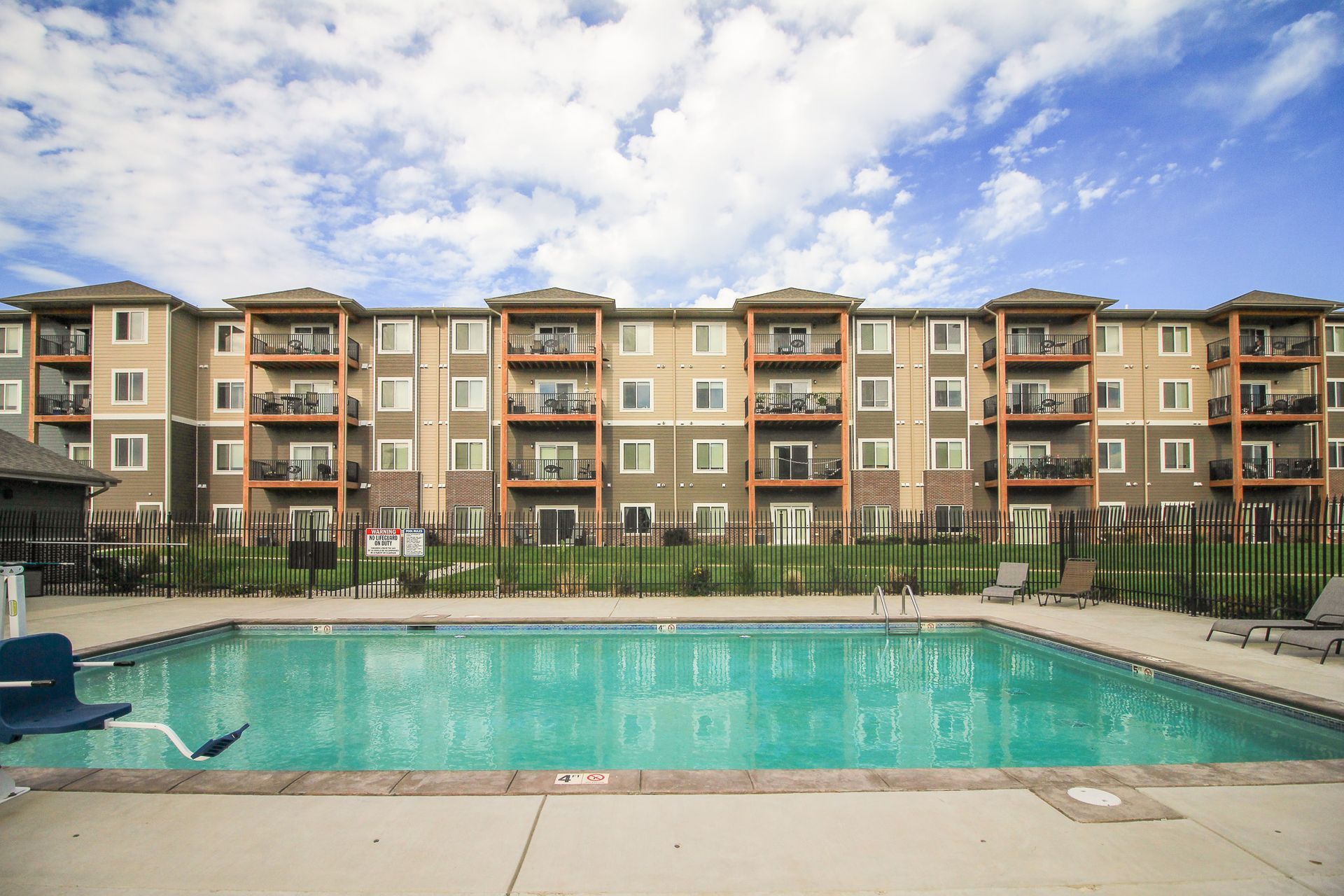 A four-story apartment building with balconies overlooks a swimming pool. The sky is blue with scattered clouds.