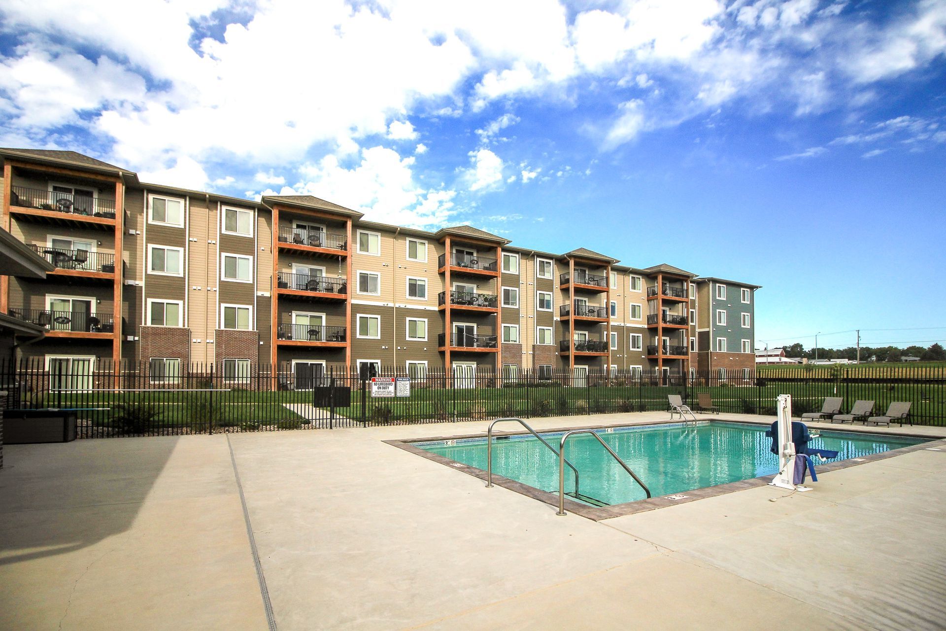 Apartment building with a pool on a sunny day. The building has multiple balconies, various colors, and a small fence.