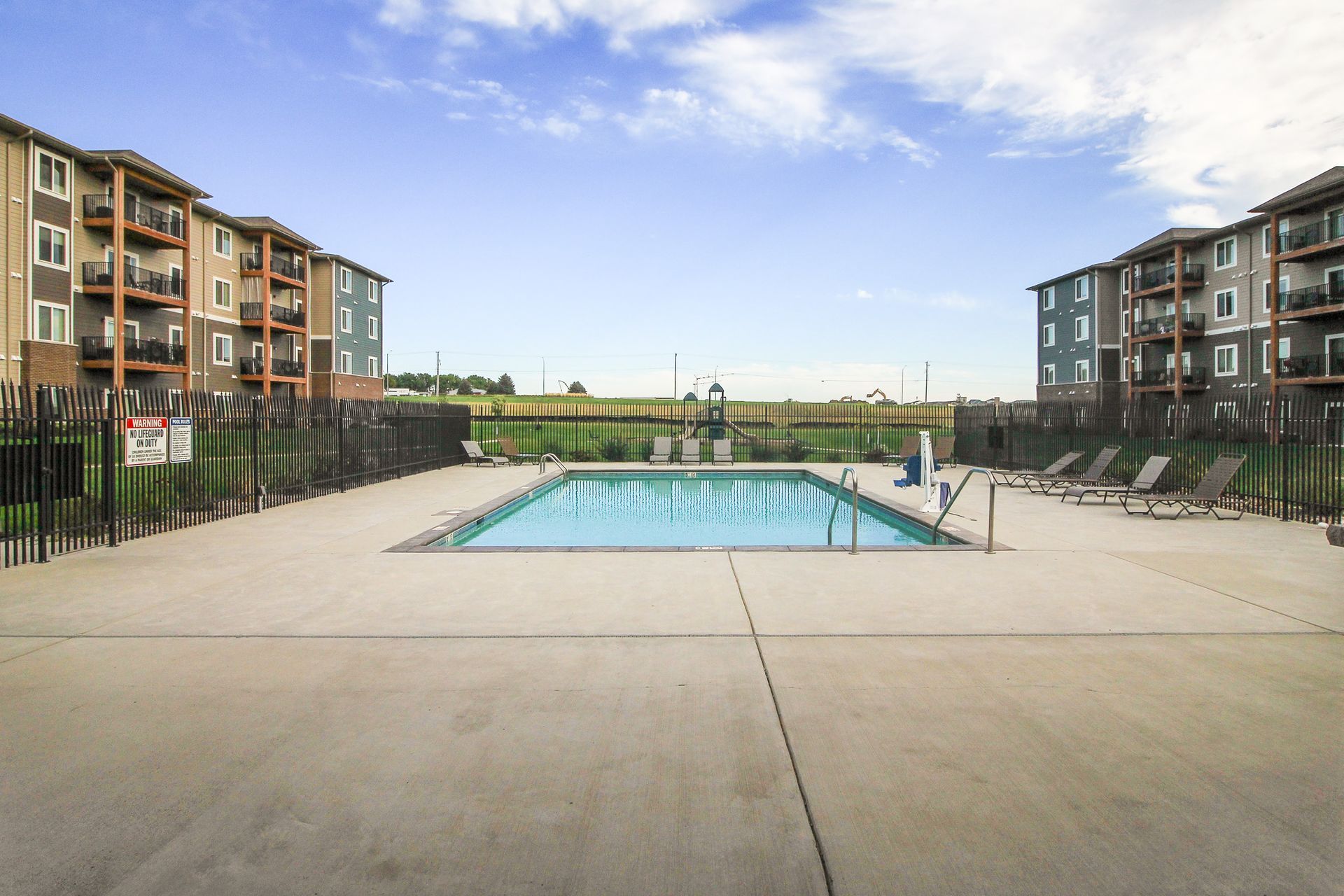 A swimming pool is surrounded by a concrete patio and fenced off from two apartment buildings under a blue sky.