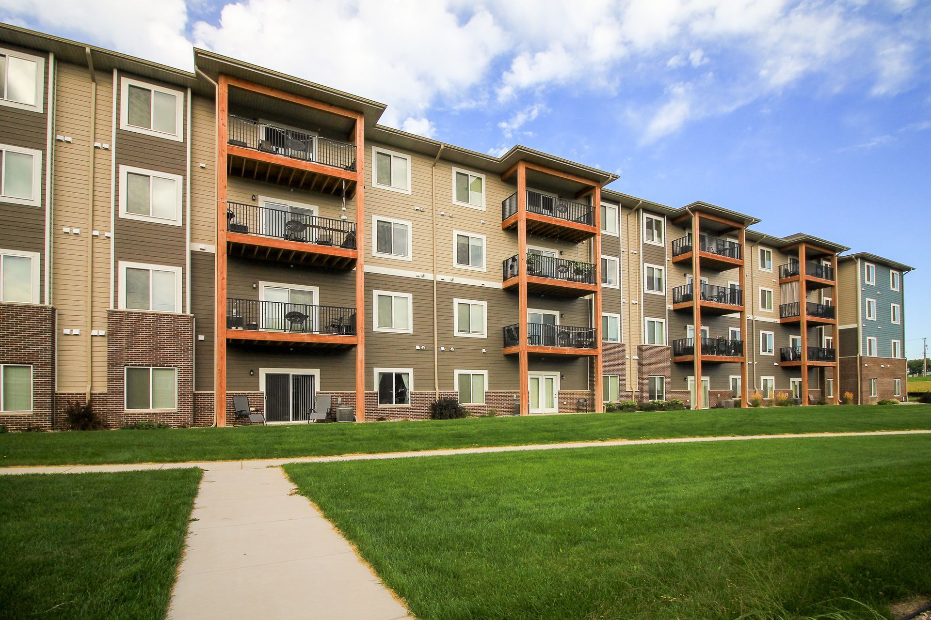 Multi-story apartment building with balconies, set against a blue sky and green lawn. Exterior features various shades of tan and blue.