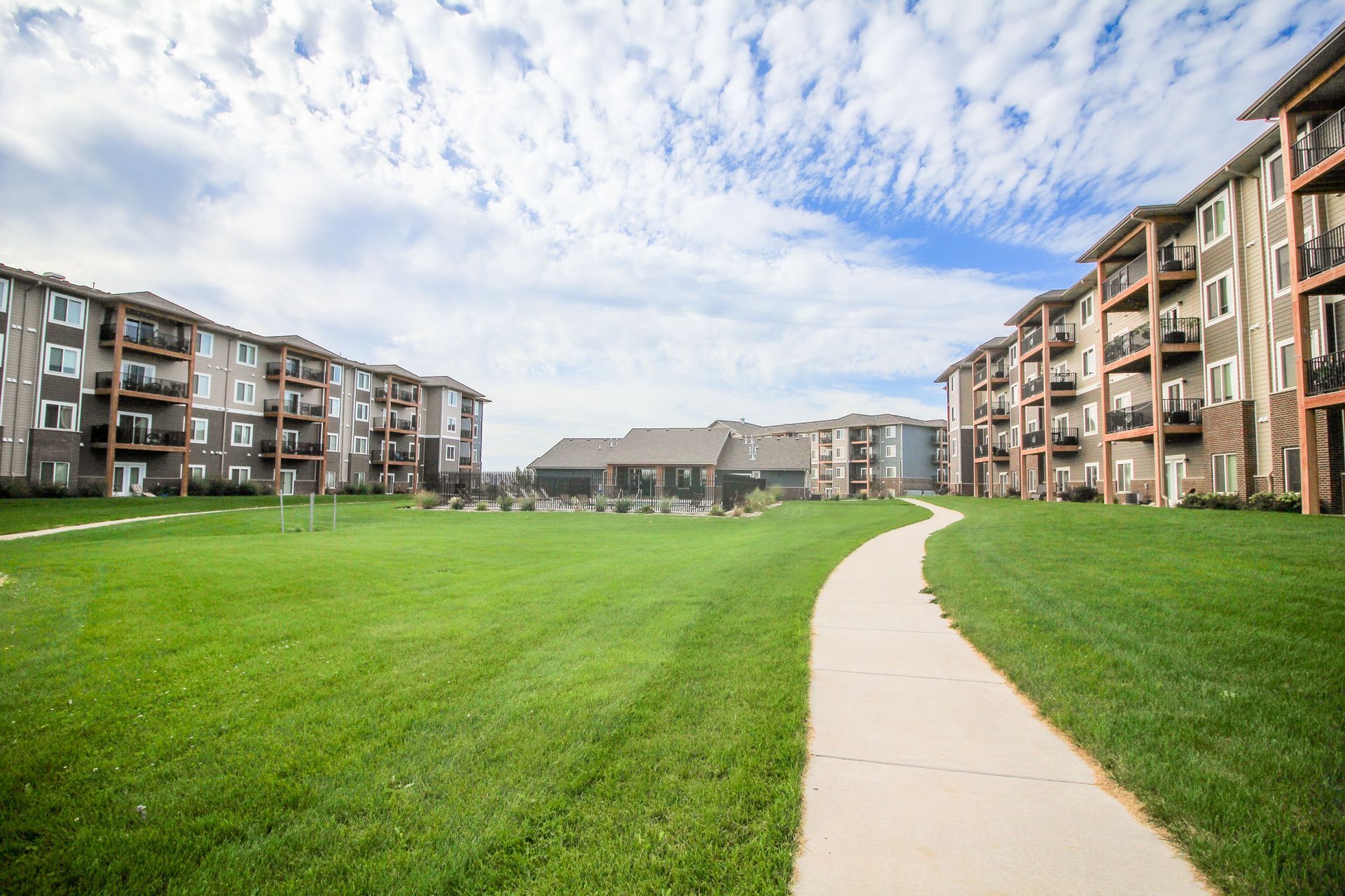 Apartment complex with green lawn, paved path, and partly cloudy sky. Buildings are multi-story with balconies.