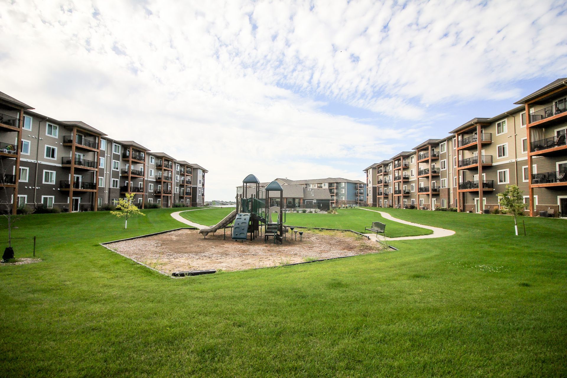 A park with a playground, surrounded by apartment buildings and a grassy lawn under a cloudy sky.