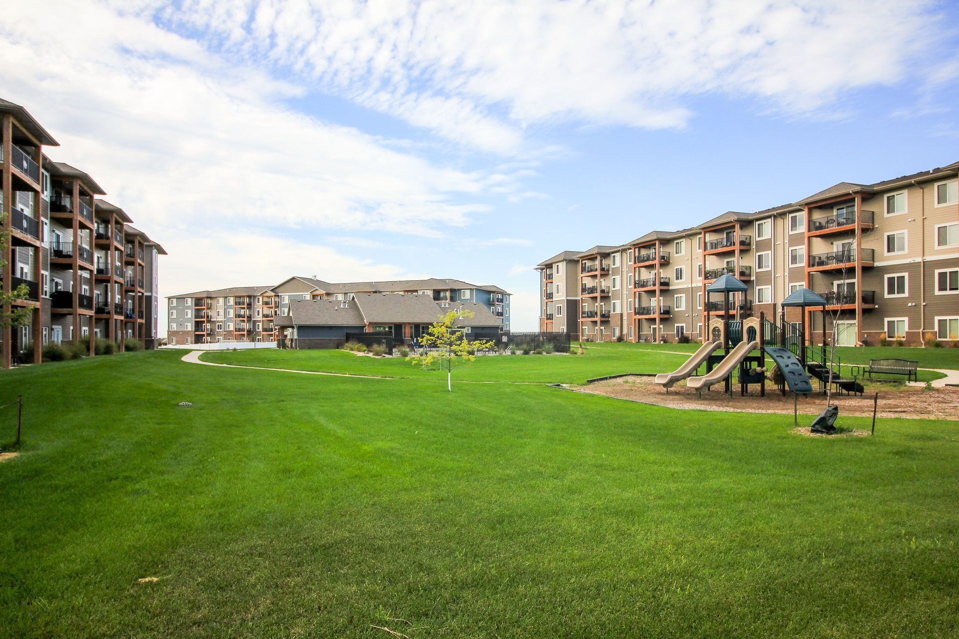Apartment complex with green lawns, playground, and buildings under a partly cloudy sky.