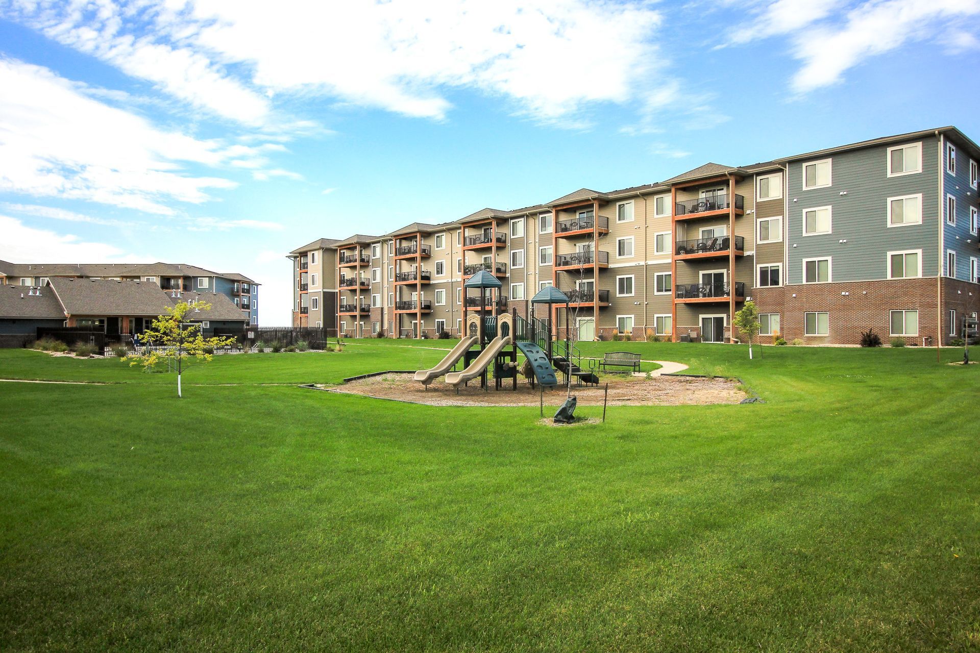 A playground in a grassy area with apartment buildings in the background under a blue sky.