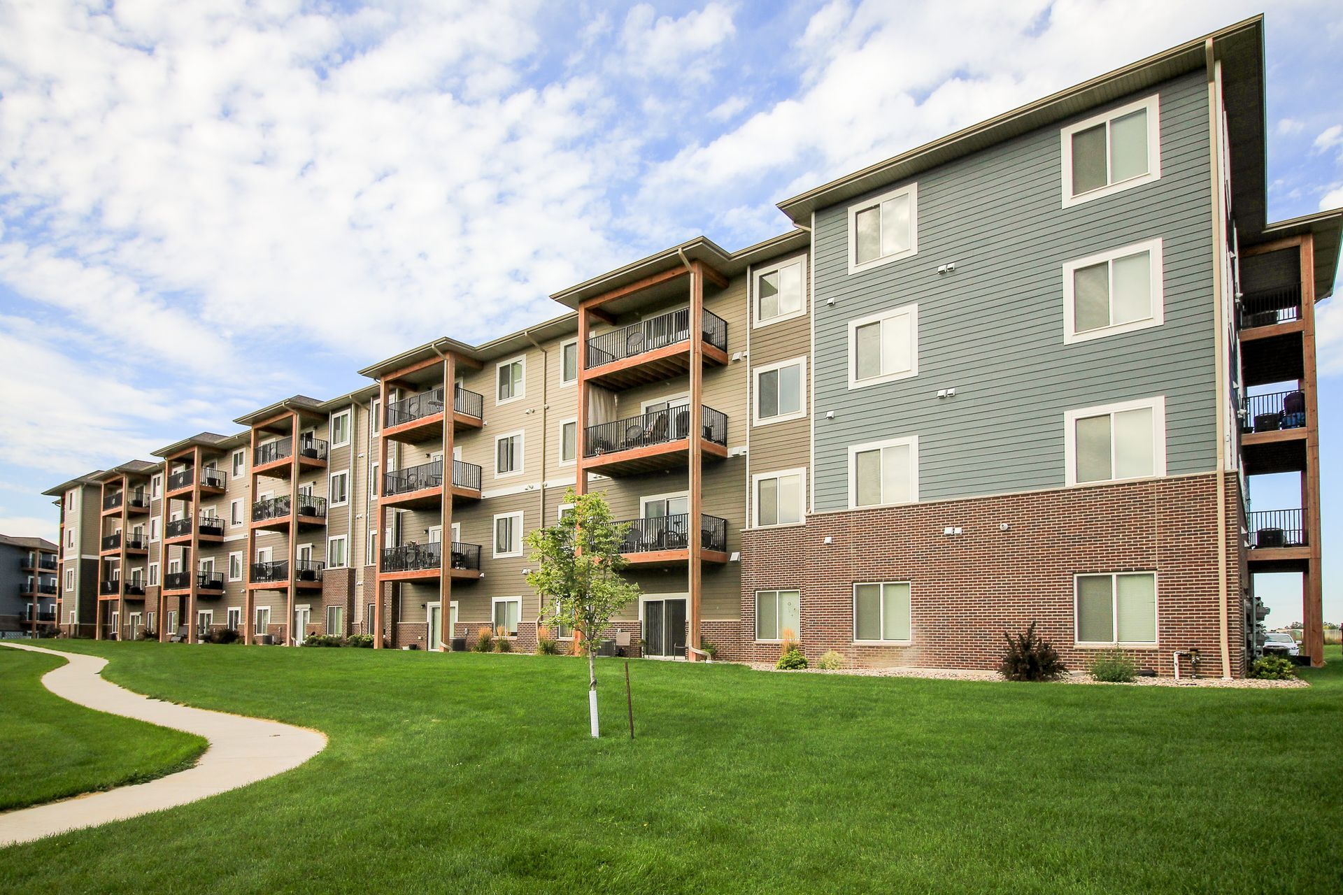 Apartment building with balconies, featuring grey and tan siding, set against a green lawn and a cloudy sky.