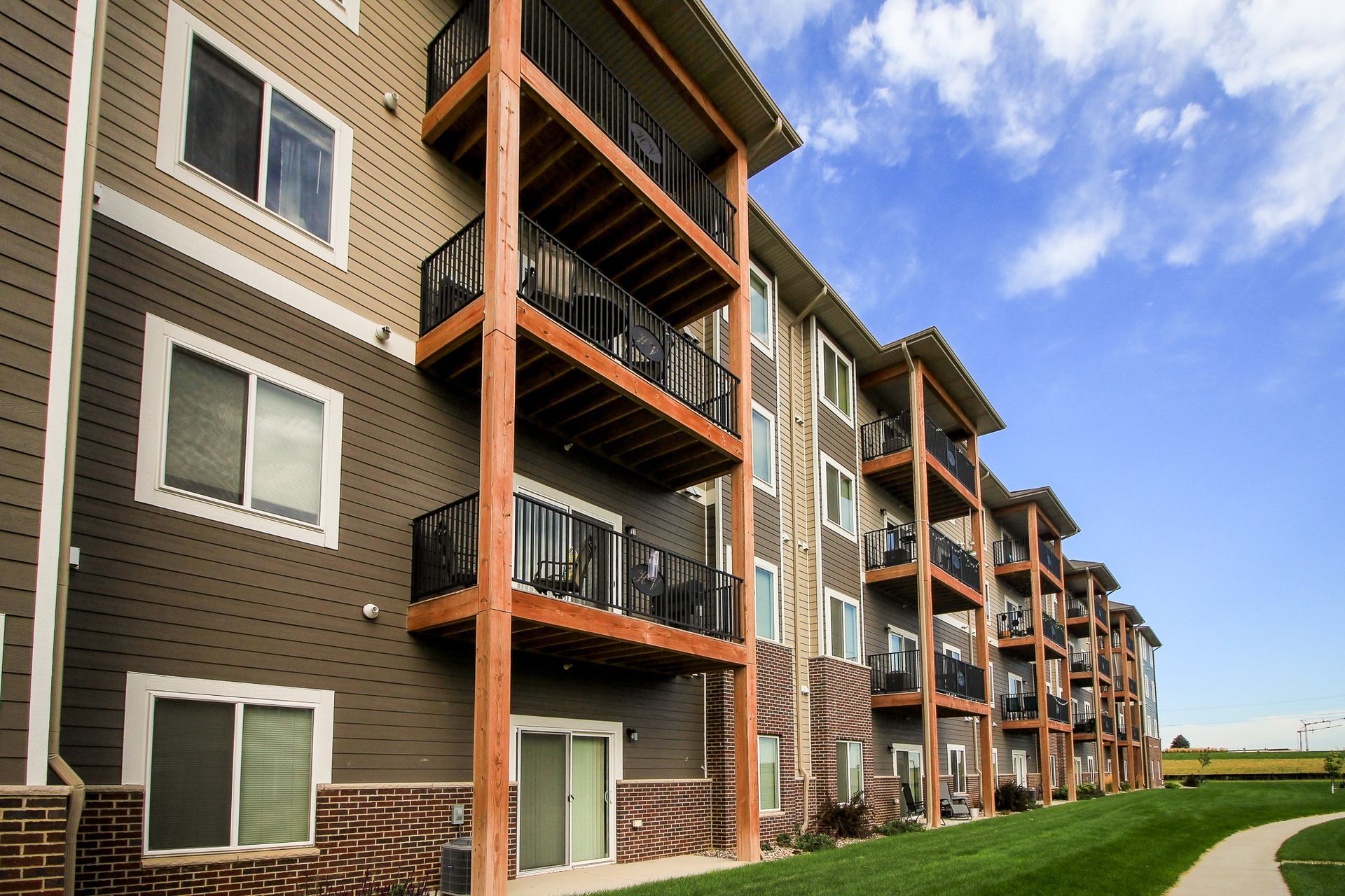 Apartment building exterior with multiple stories, brown siding, balconies, and a blue sky with clouds.