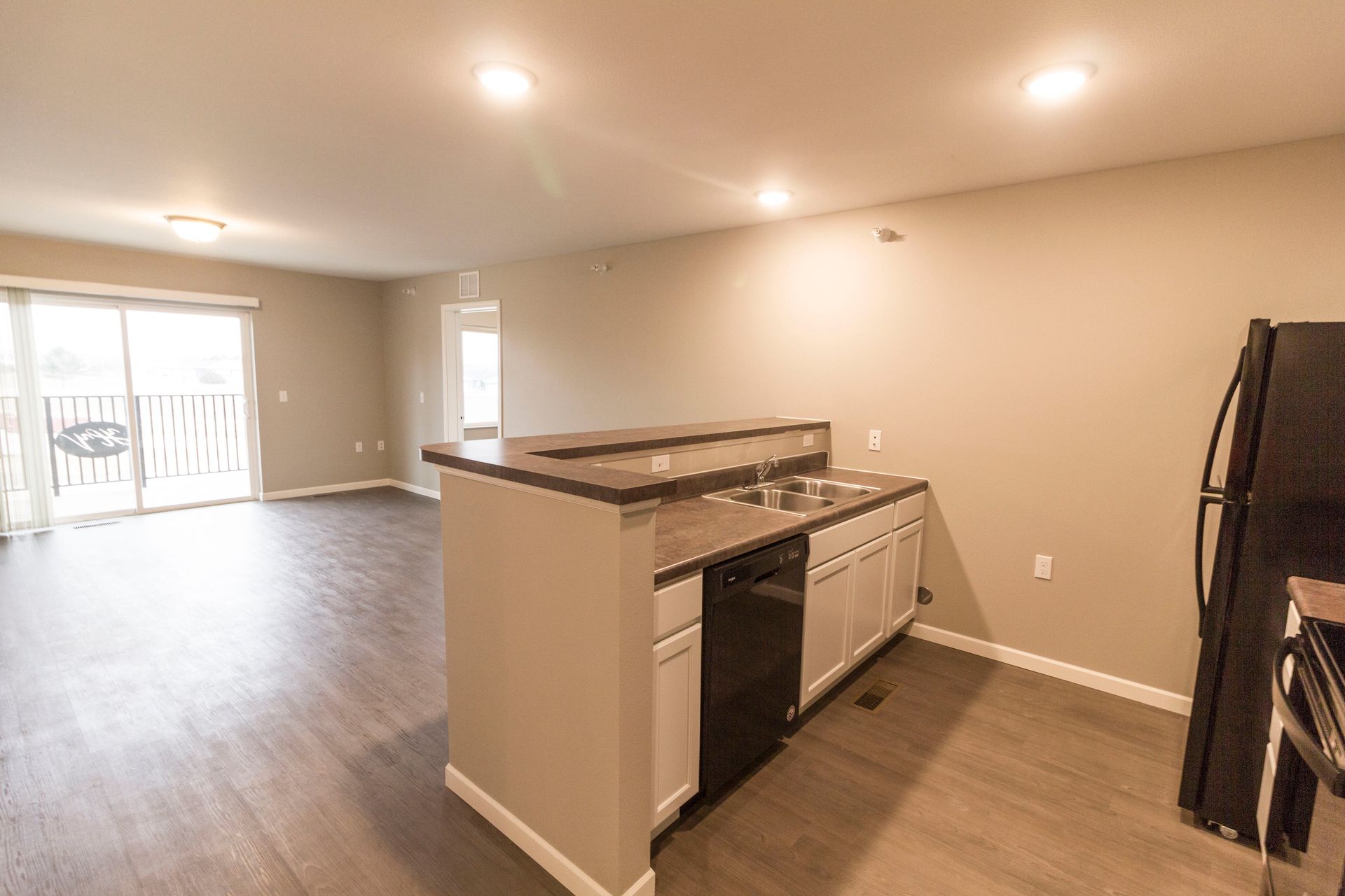 Open-concept apartment kitchen with a dark countertop island with a sink and dishwasher, and a black refrigerator. The room has hardwood floors, beige walls, and a glass door leading to a balcony.