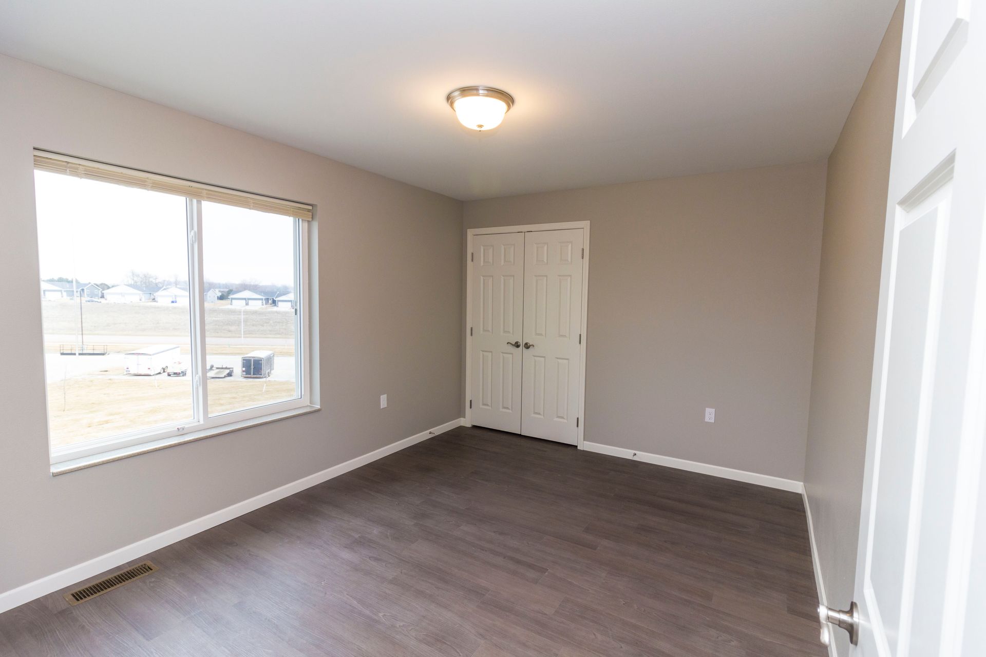 Empty bedroom with gray walls, wood flooring, and a large window overlooking a neighborhood. A closed door is visible.