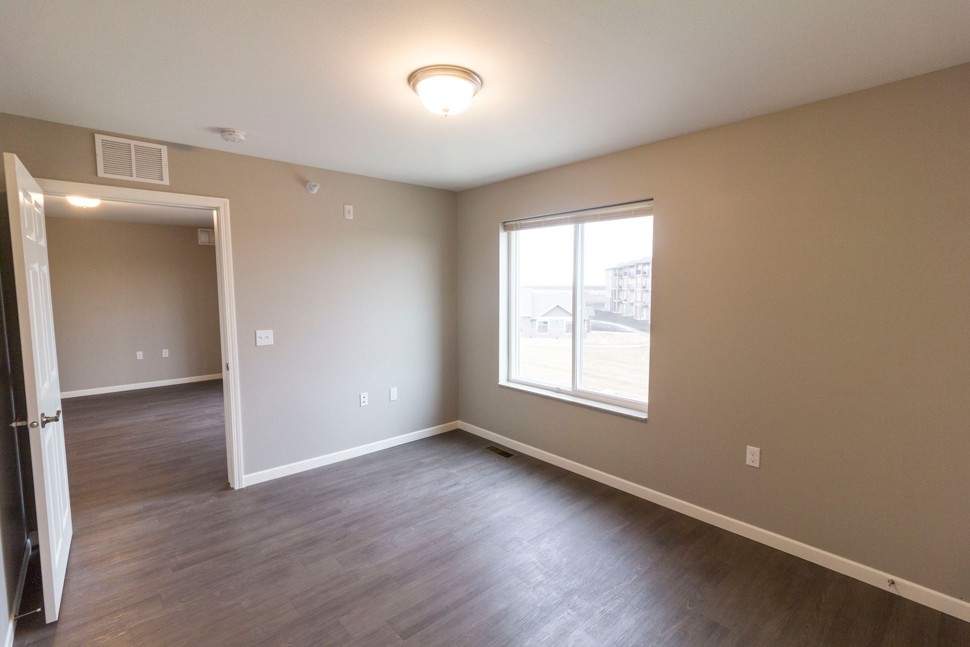 Empty bedroom with gray walls, wood-look flooring, and a window. An open door leads to another room.
