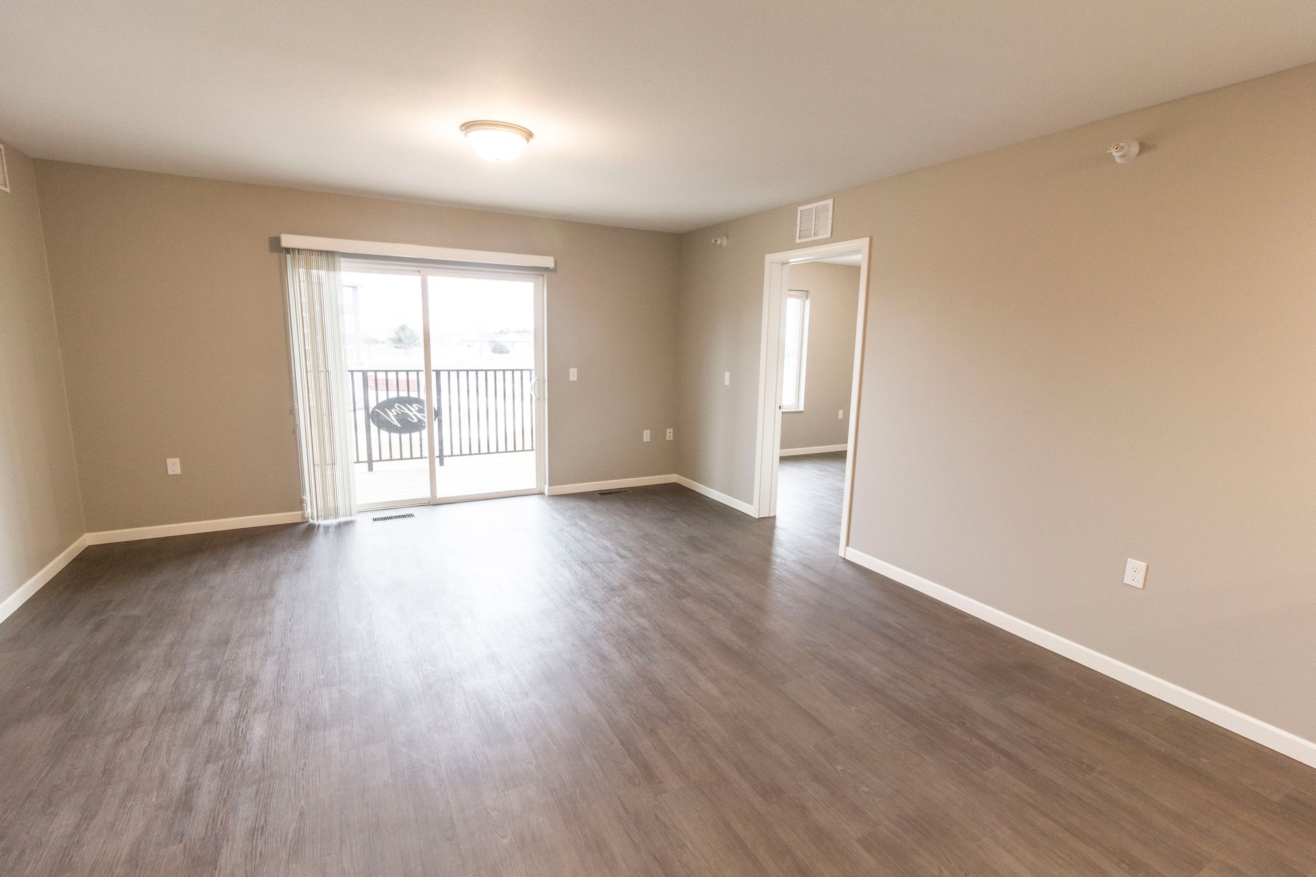 Empty living room with dark wood-look flooring, beige walls, and a sliding glass door leading to a balcony.