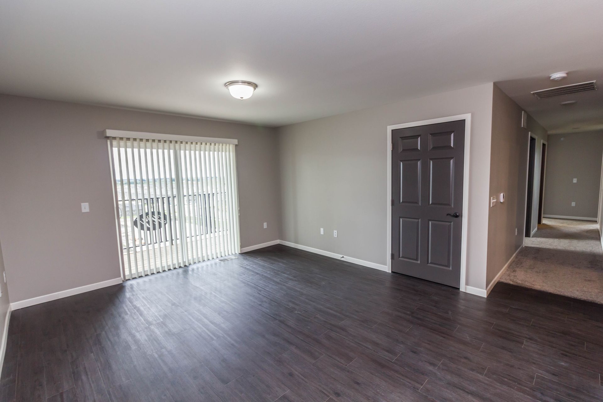 Empty living room with gray walls, dark wood flooring, and a sliding glass door covered by blinds. A dark gray door is on the right.
