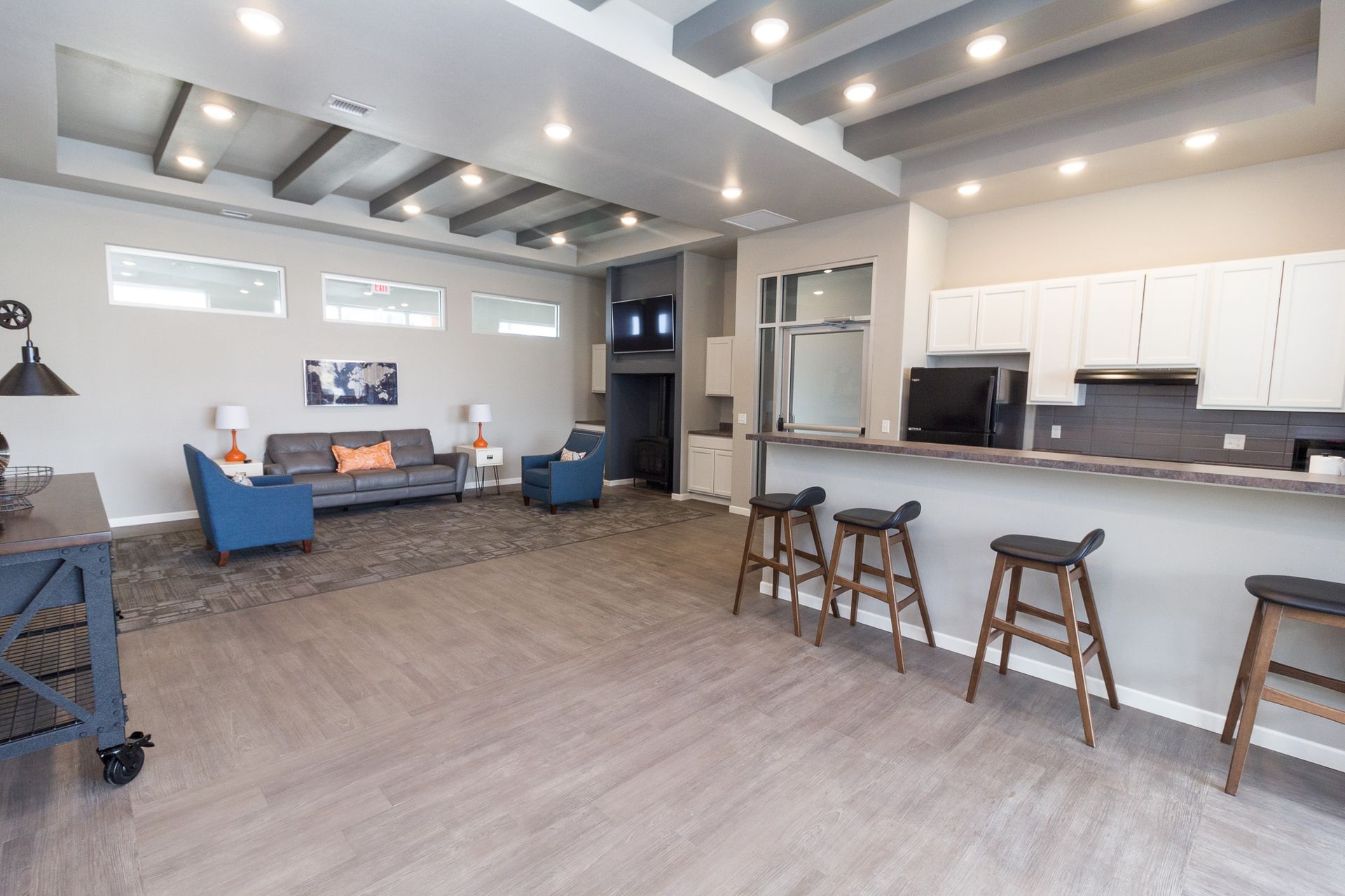Spacious community lounge with seating areas, a kitchen, and a bar with stools. Gray walls, wood floors, and a bright, modern feel.