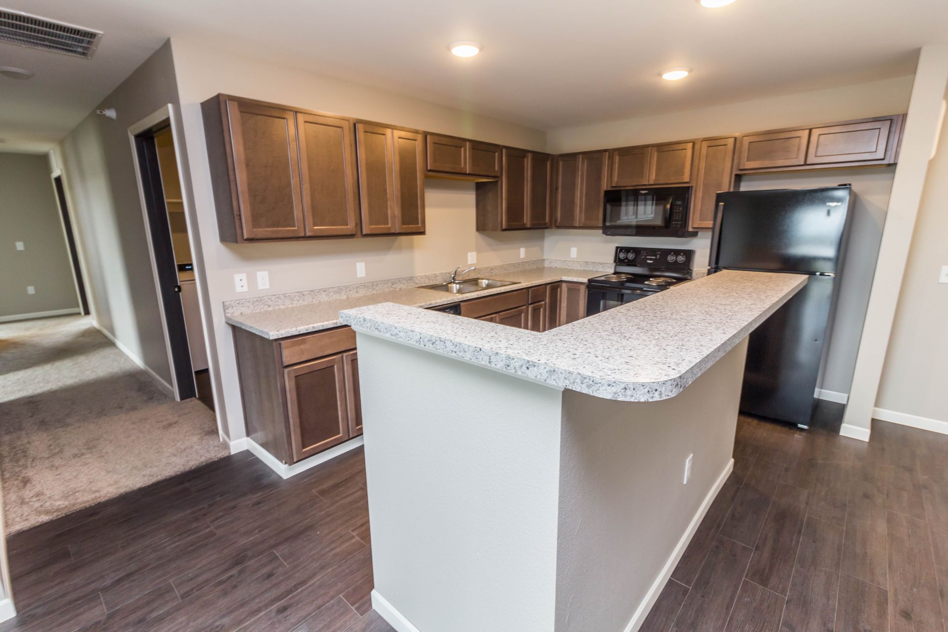 A modern kitchen with dark wood cabinets, light countertops, and a breakfast bar. Appliances are black. Dark wood flooring.