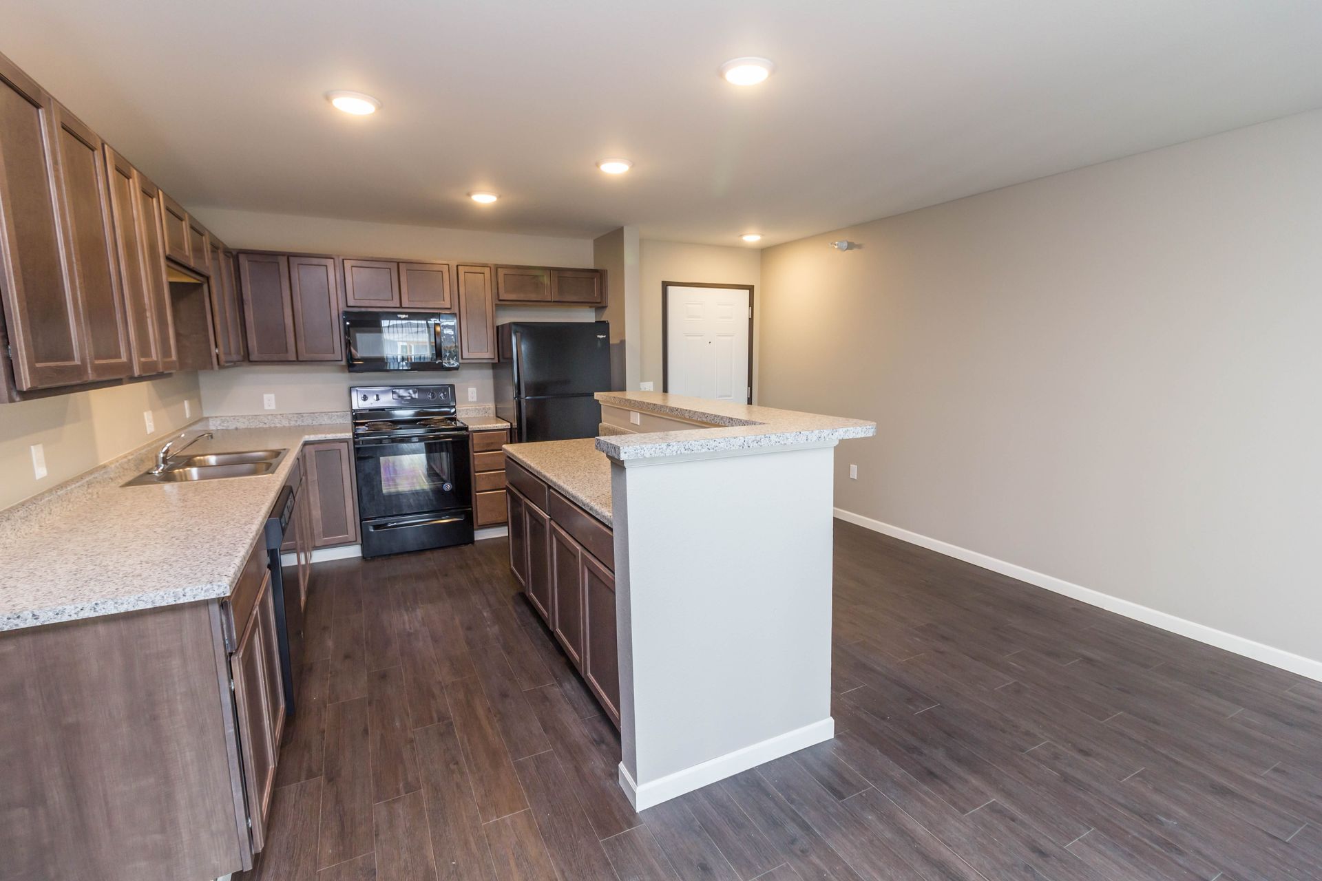 A modern kitchen with dark wood cabinets, gray countertops, and a central island, featuring black appliances and dark wood flooring.