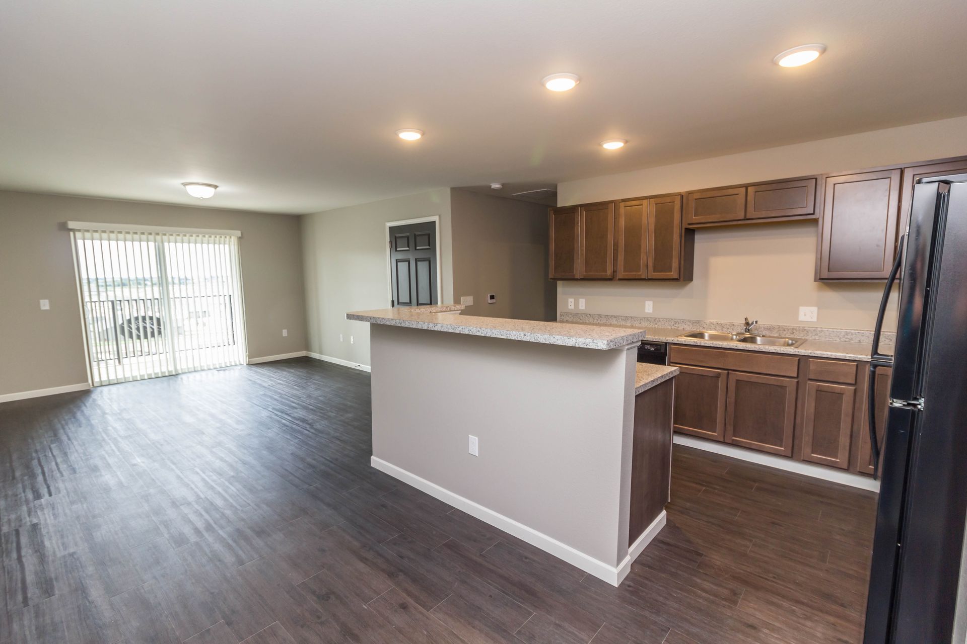 Interior view of a modern kitchen and living space with dark wood flooring, granite countertops, and brown cabinets. A sliding glass door leads to an outdoor area.