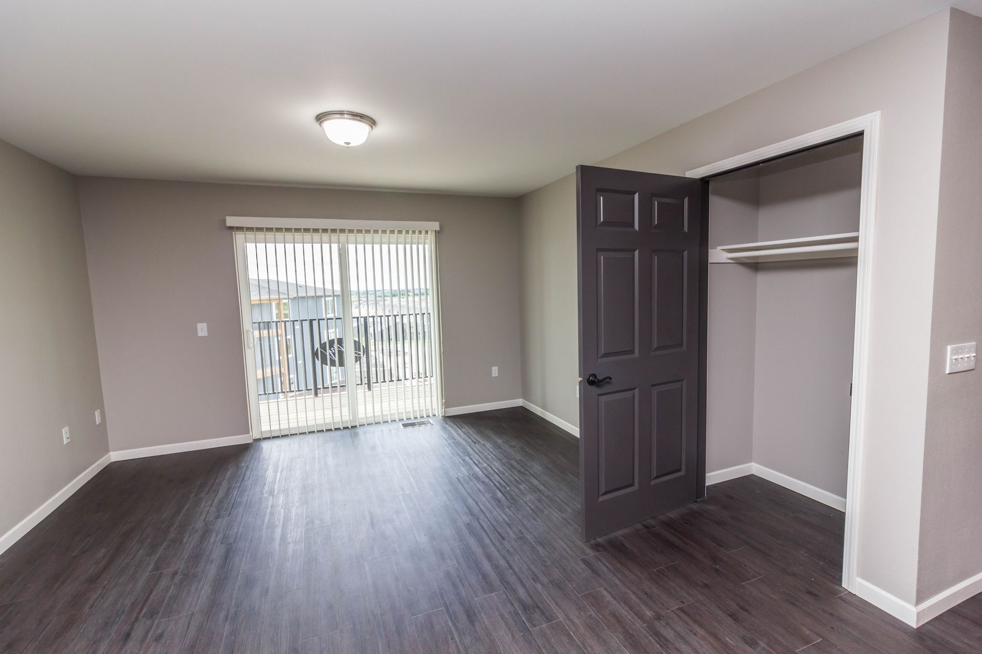 Empty bedroom with dark wood floors, sliding glass door, and open closet. Dark grey door stands ajar.