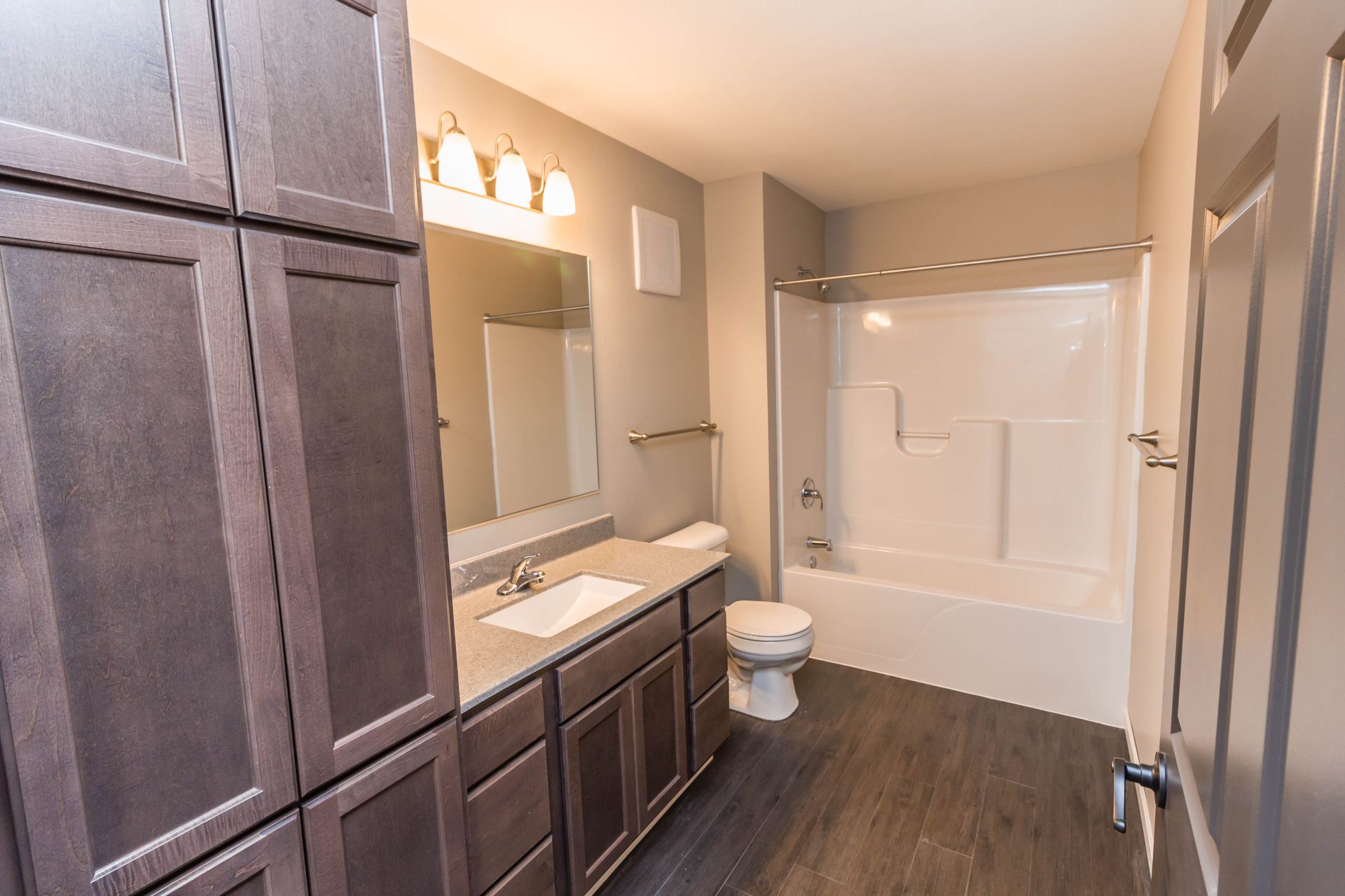 A modern bathroom with dark wood cabinets and flooring, a white tub/shower, and a white toilet.