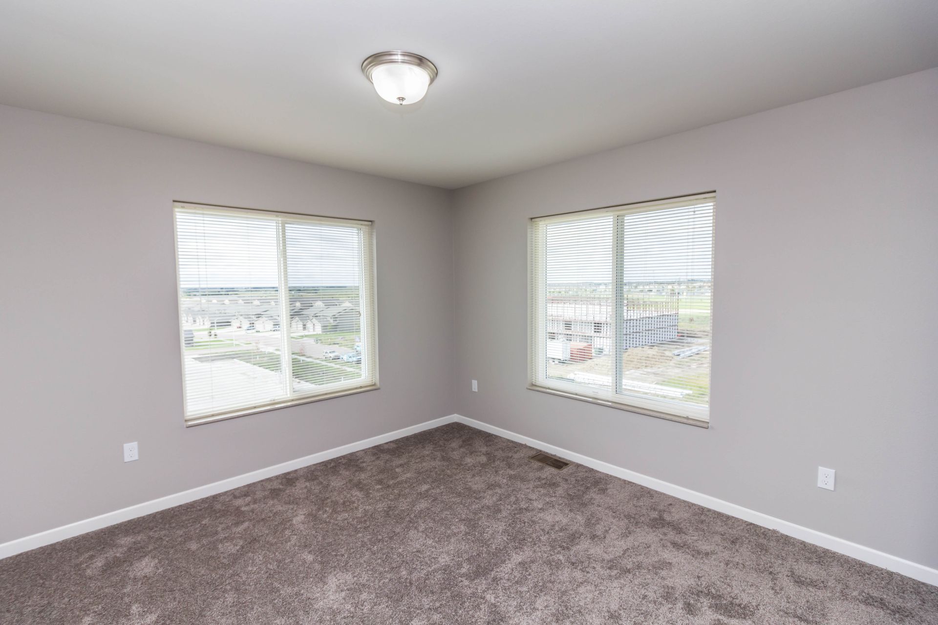 Empty bedroom with gray carpet, two windows, and a ceiling light. Walls are painted light gray.