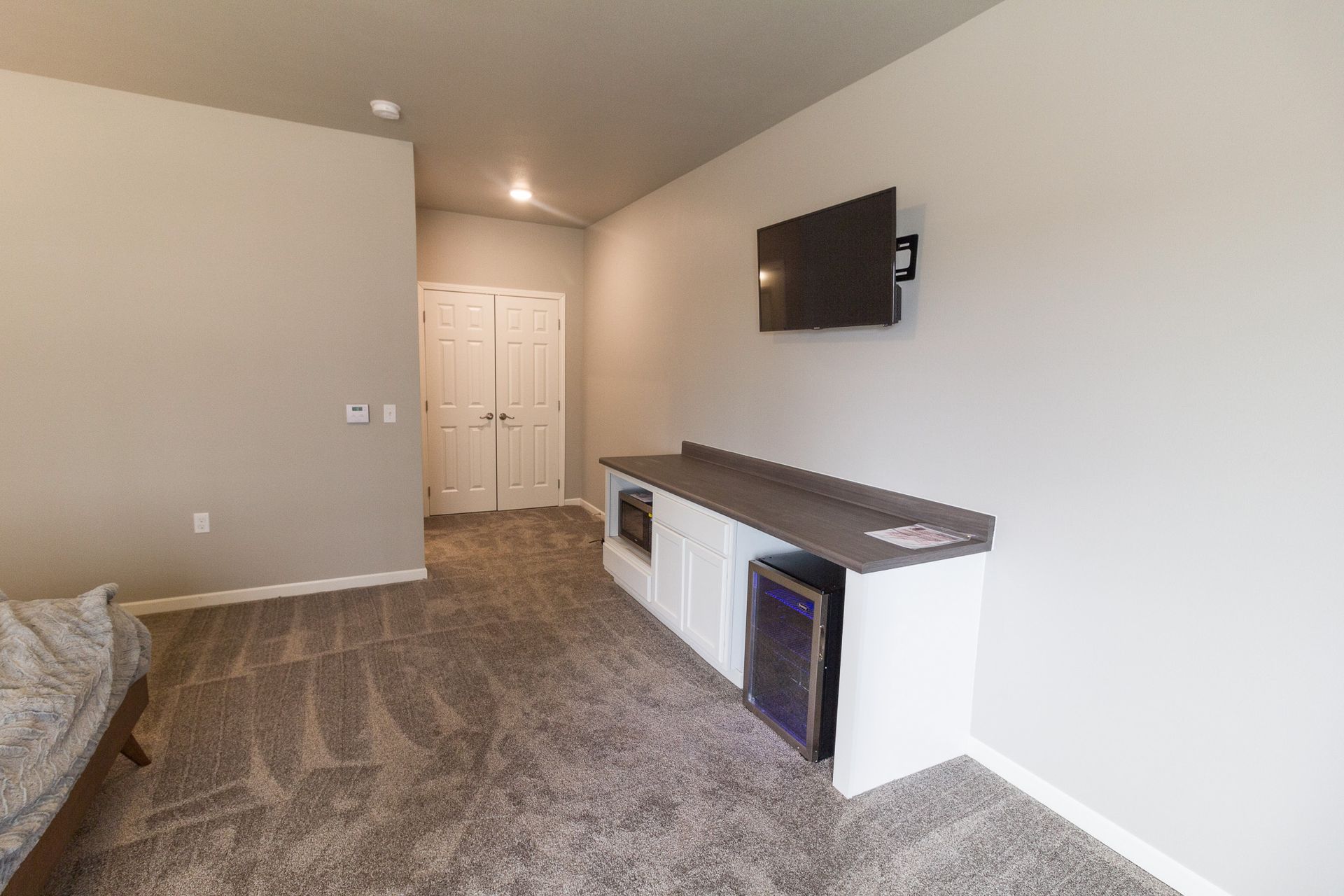 A hotel room with a white cabinet, a microwave, and a mini-fridge, a TV mounted on the wall, and a closed door in the background.