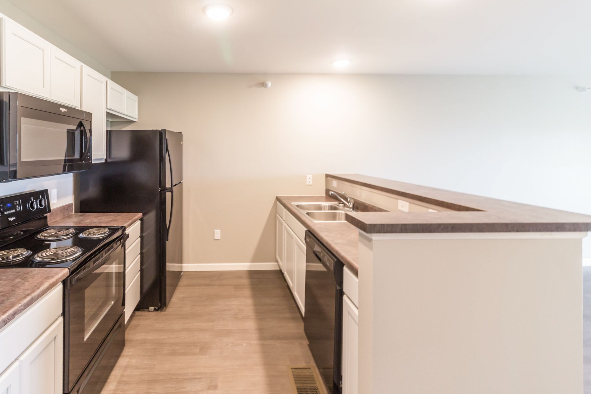 A kitchen with white cabinets, black appliances, and a countertop bar. The walls are light gray with wood-look flooring.