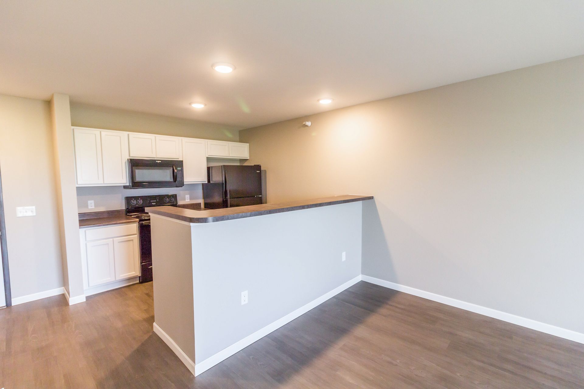 Interior view of a modern kitchen and living area. It features white cabinets, black appliances, a breakfast bar, and wood-look flooring.
