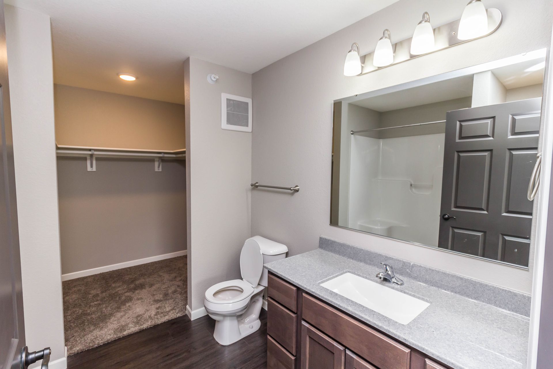 Bathroom with dark wood cabinets, a gray countertop, a white toilet, and a shower visible in the mirror. A closet is in the background.