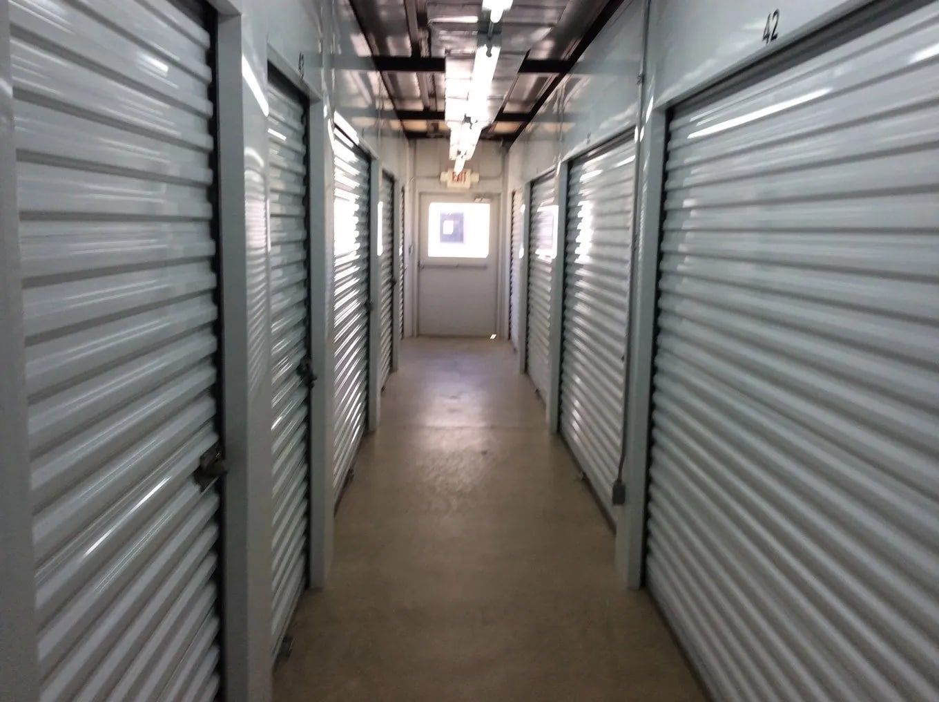 A long, narrow hallway lined with closed, metal storage unit doors. Bright lights shine overhead, a door at the end.